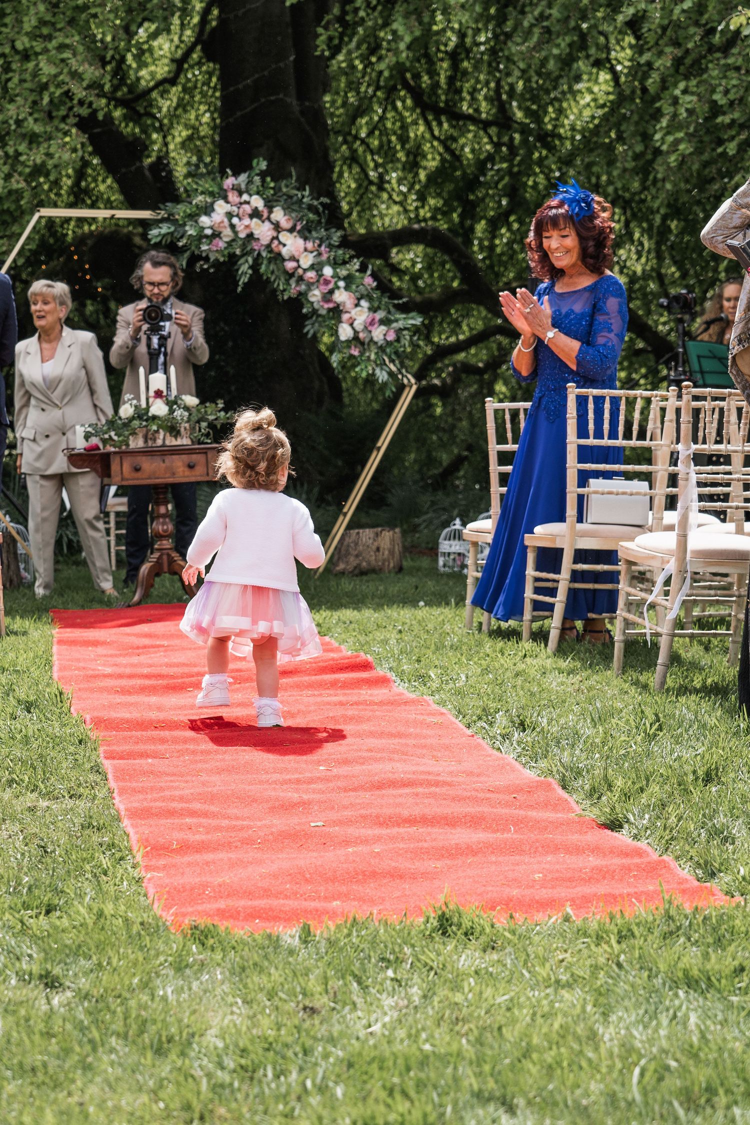 Curly-haired flower girl in white dress walks down red carpet aisle at elegant outdoor garden wedding ceremony.
