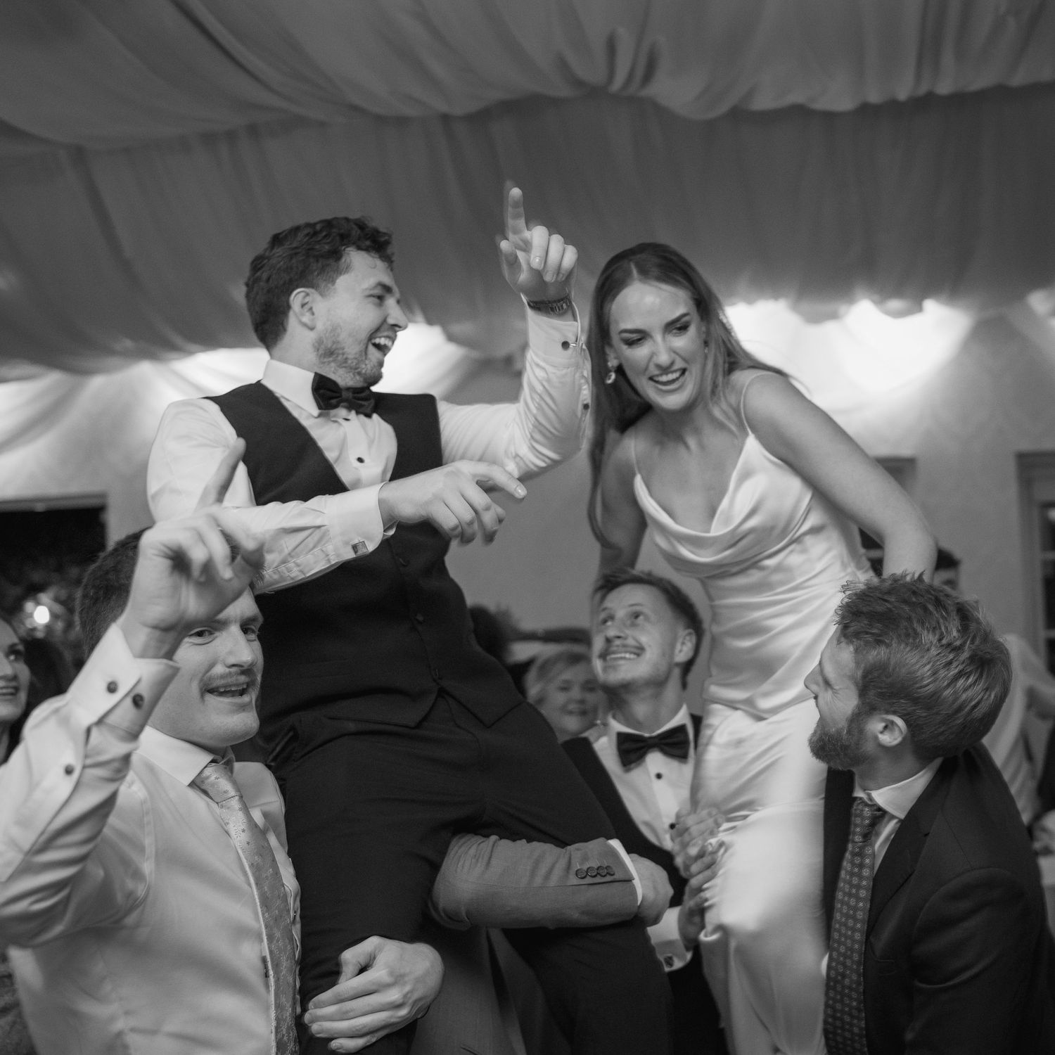 Bride and groom lifted on shoulders by groomsmen during lively wedding reception dance.