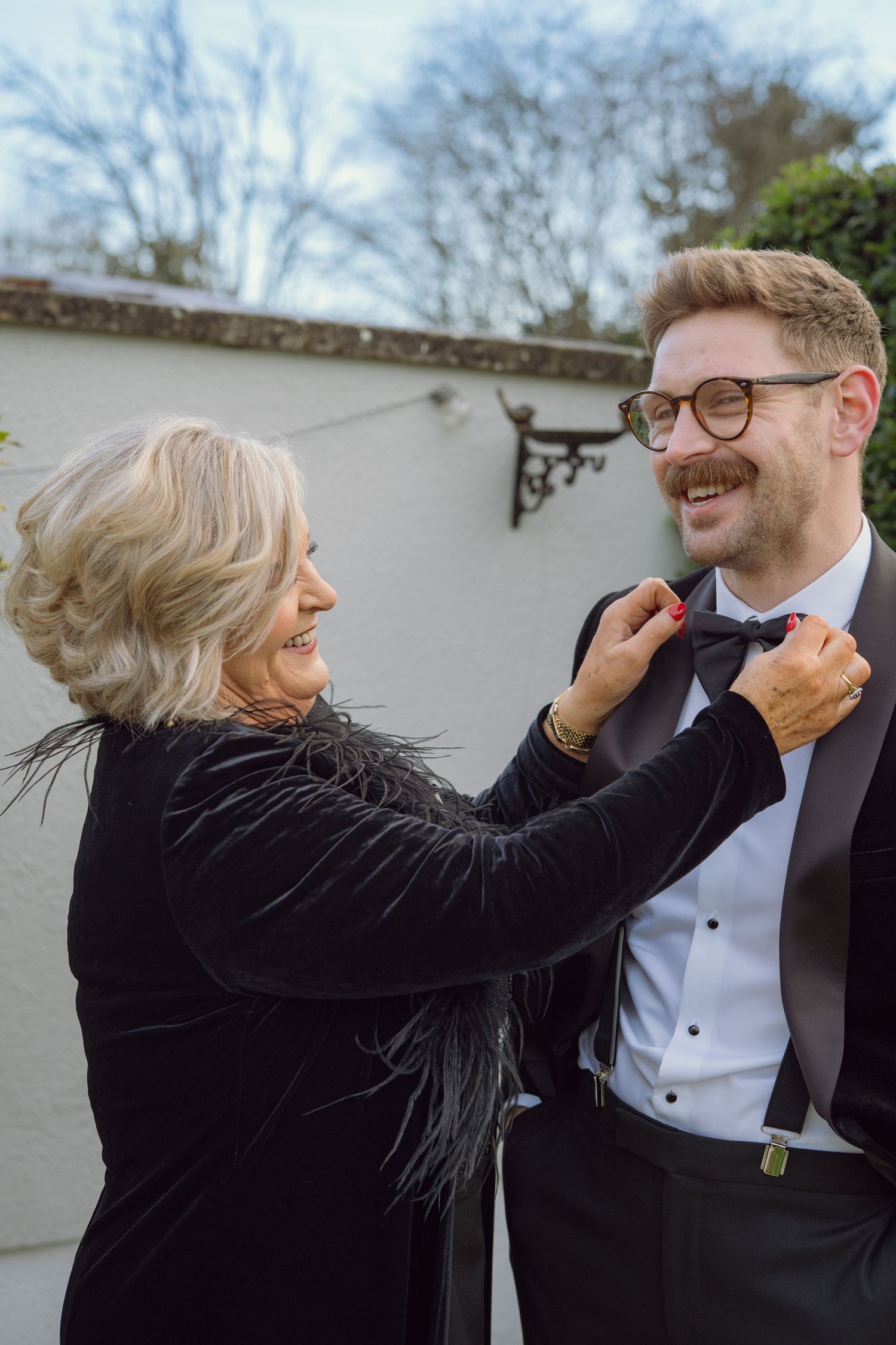 Mother adjusts groom's bow tie outdoors, both smiling, in elegant wedding attire.