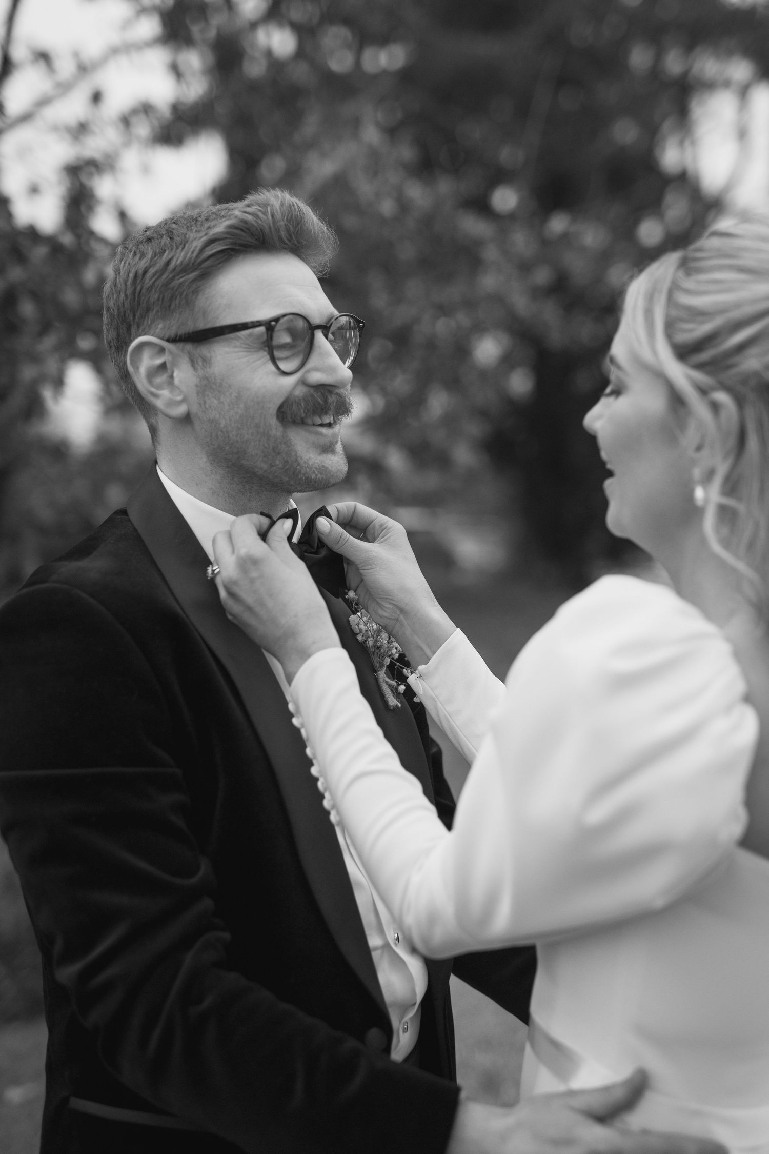 Black and white photo of bride adjusting groom's bow tie outdoors at wedding.