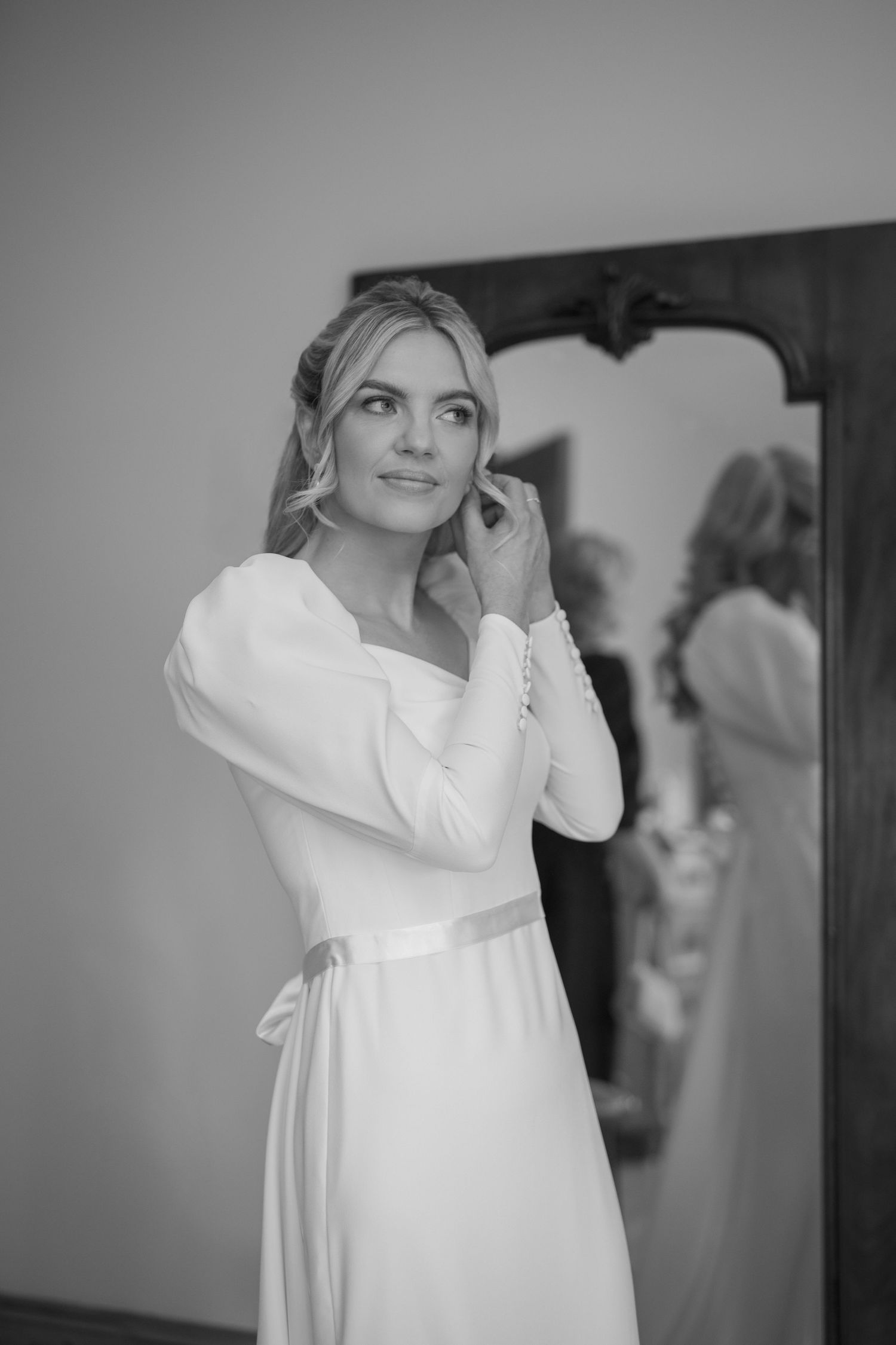 Elegant bride in long-sleeve white gown adjusts earring before mirror in black and white portrait.