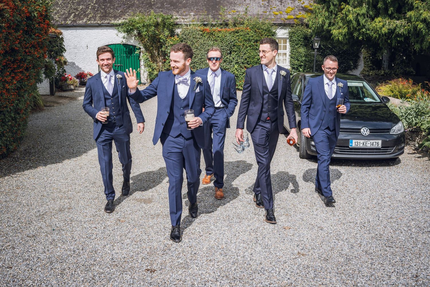 Groom and four groomsmen in matching navy suits walking confidently across a gravel driveway.