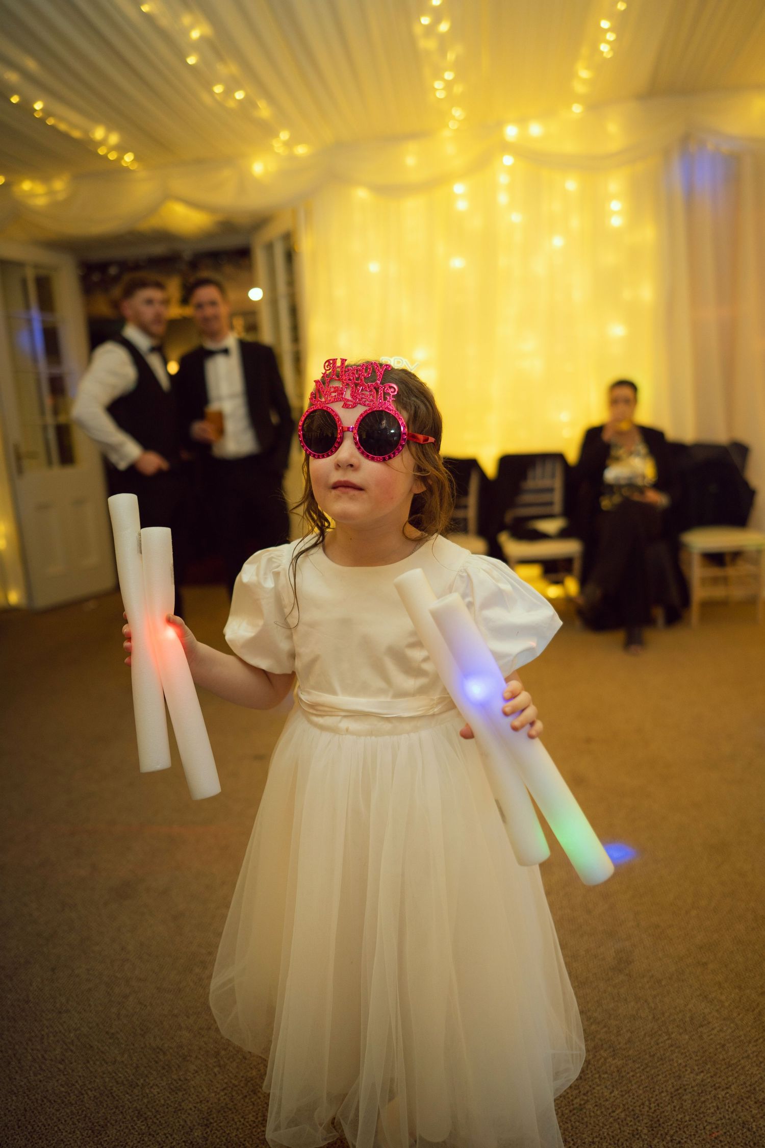 Young flower girl in white dress wearing pink novelty sunglasses holding colorful glow sticks at wedding party.