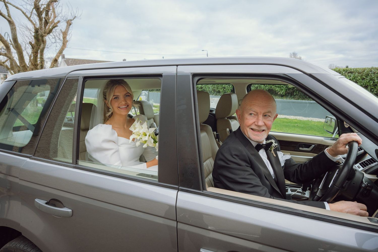 Smiling bride in white dress sitting beside elderly man in tuxedo driving silver SUV.