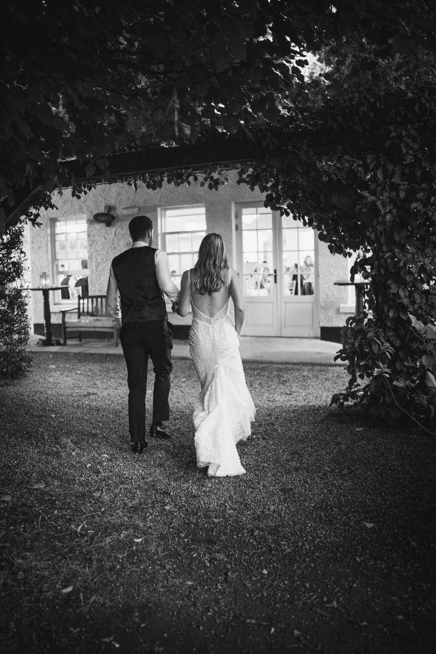 Black and white photo of a bride and groom walking hand in hand at their wedding through a vine-covered archway toward Rathsallagh House
