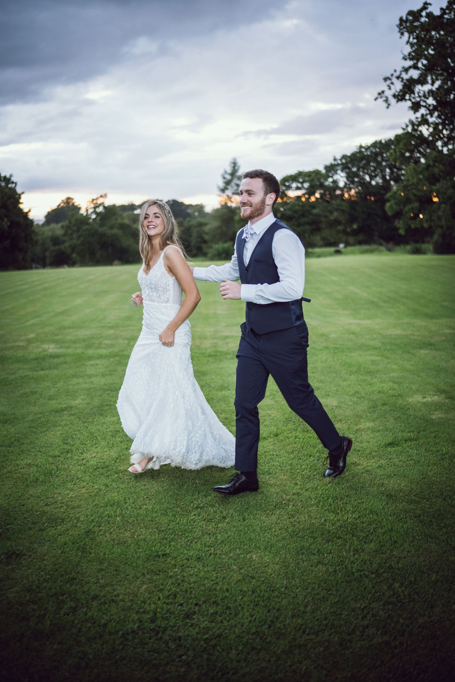 Bride in white lace gown and groom in navy vest walk joyfully across green lawn at outdoor wedding.