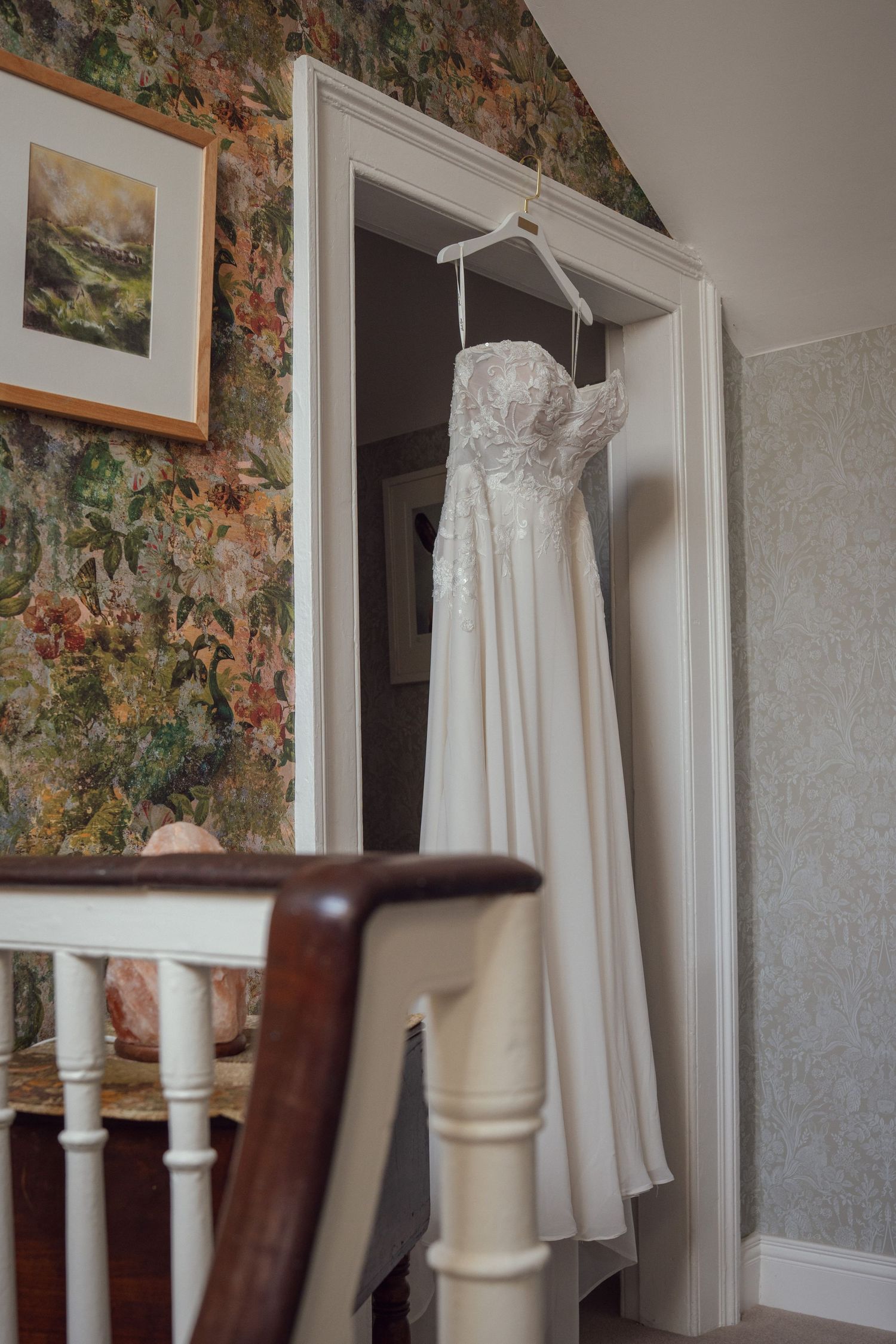 Strapless lace wedding dress hanging in a doorway of a charming floral wallpapered room.