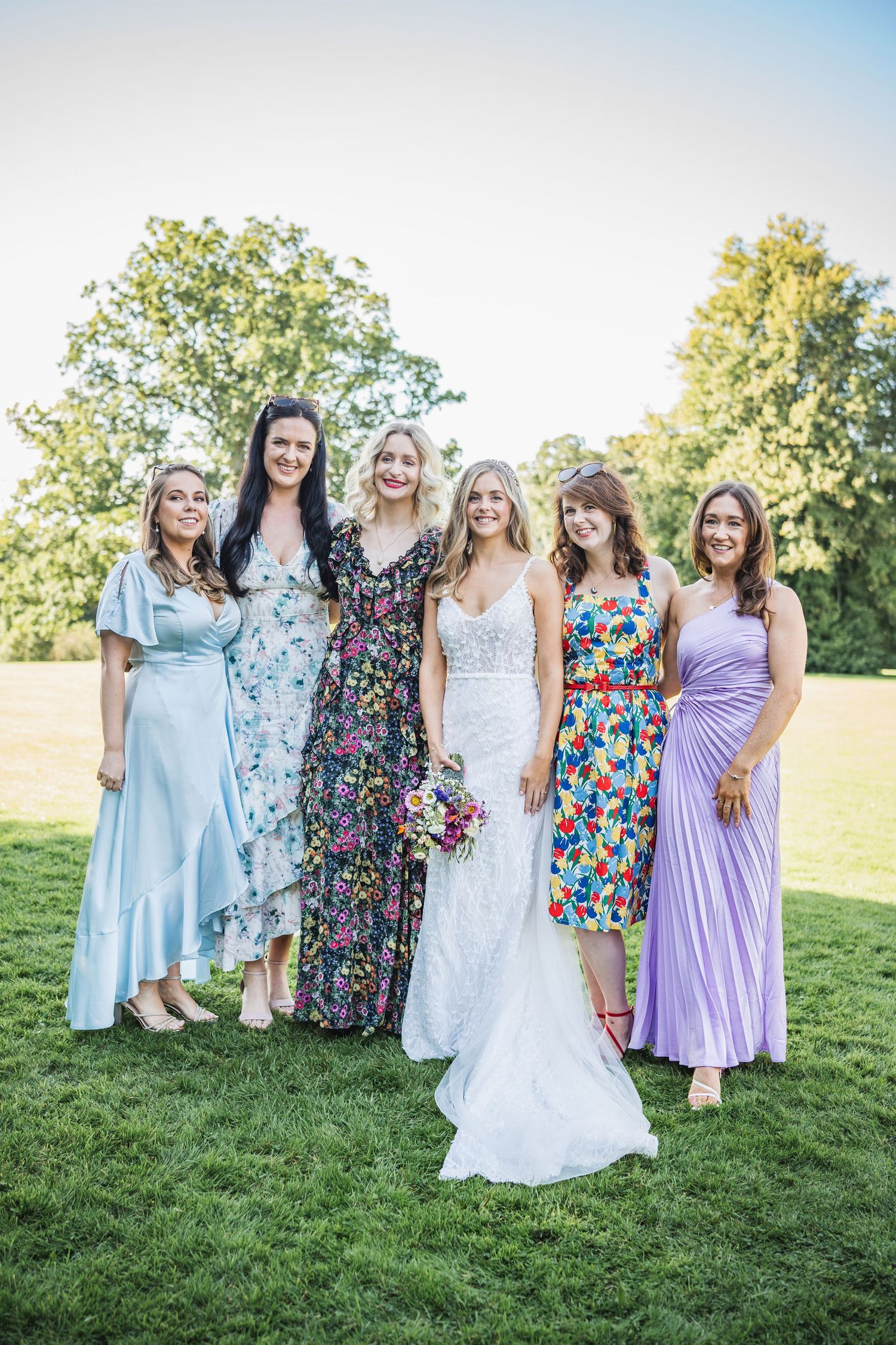 Bride in white lace gown poses with four bridesmaids in colorful dresses on green lawn at wedding.