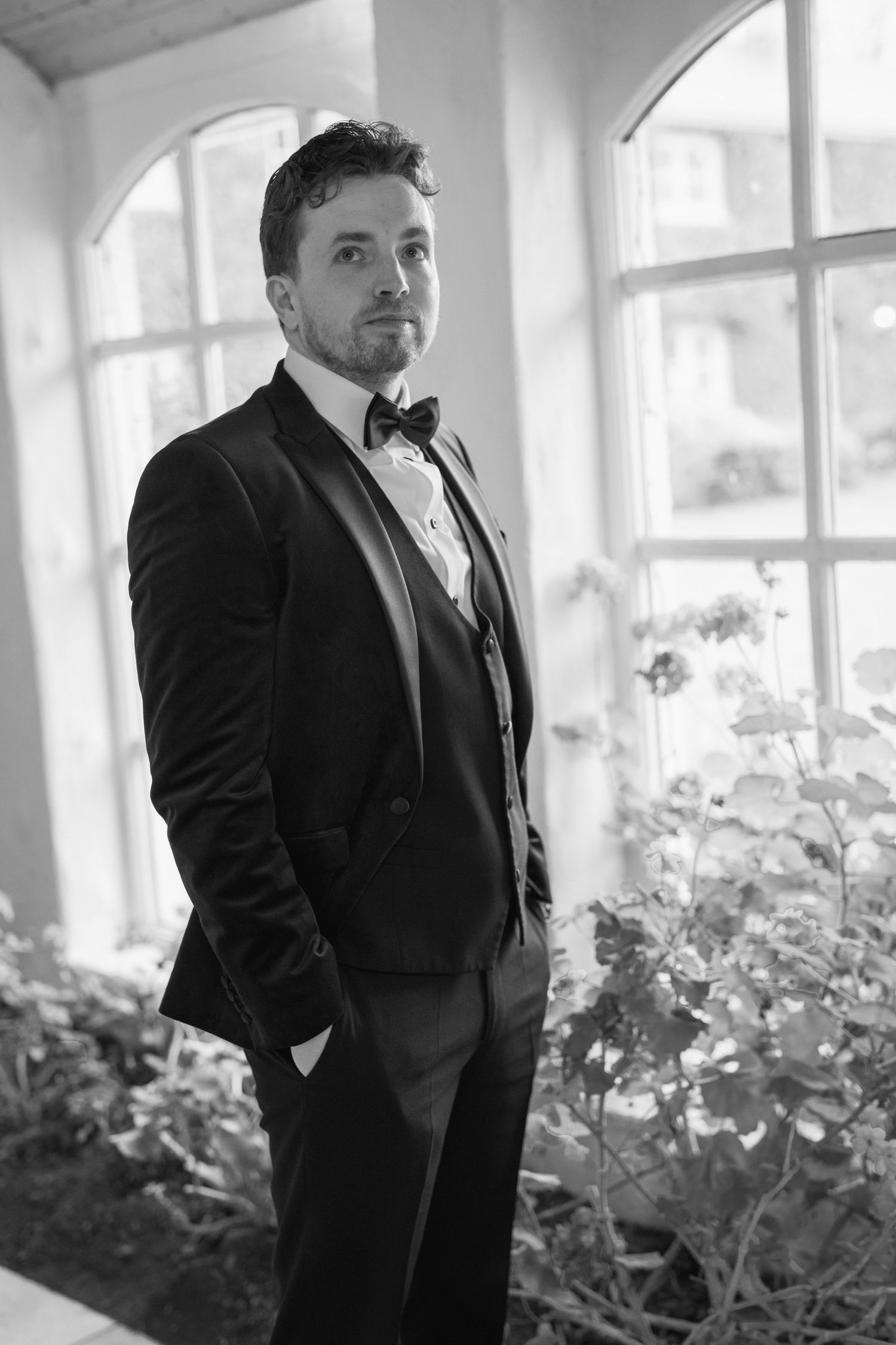 Groom in black tuxedo with bow tie posing by large windows.