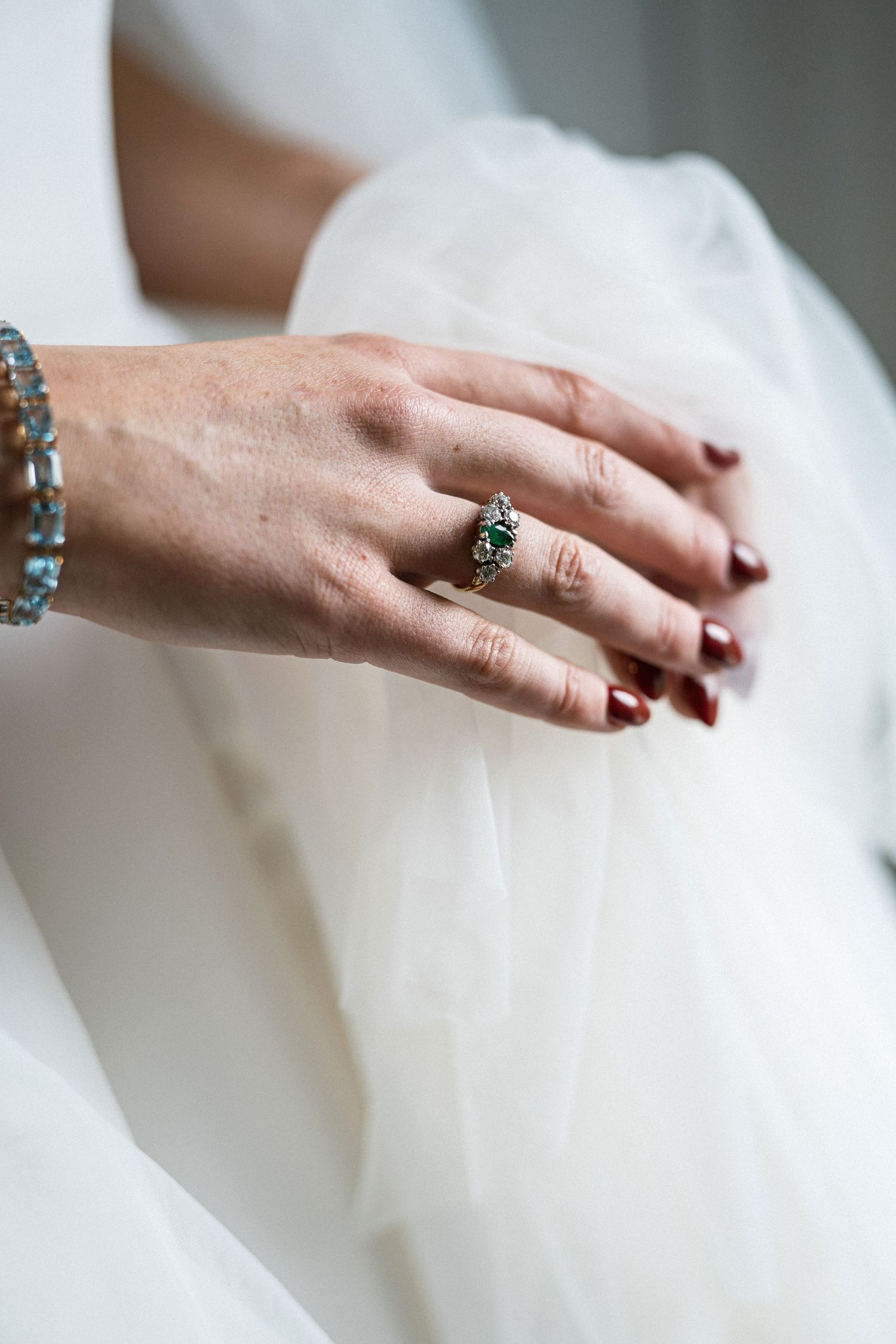 Close-up of bride's hand wearing a vintage-style teal gemstone ring with dark red nails against white wedding dress.