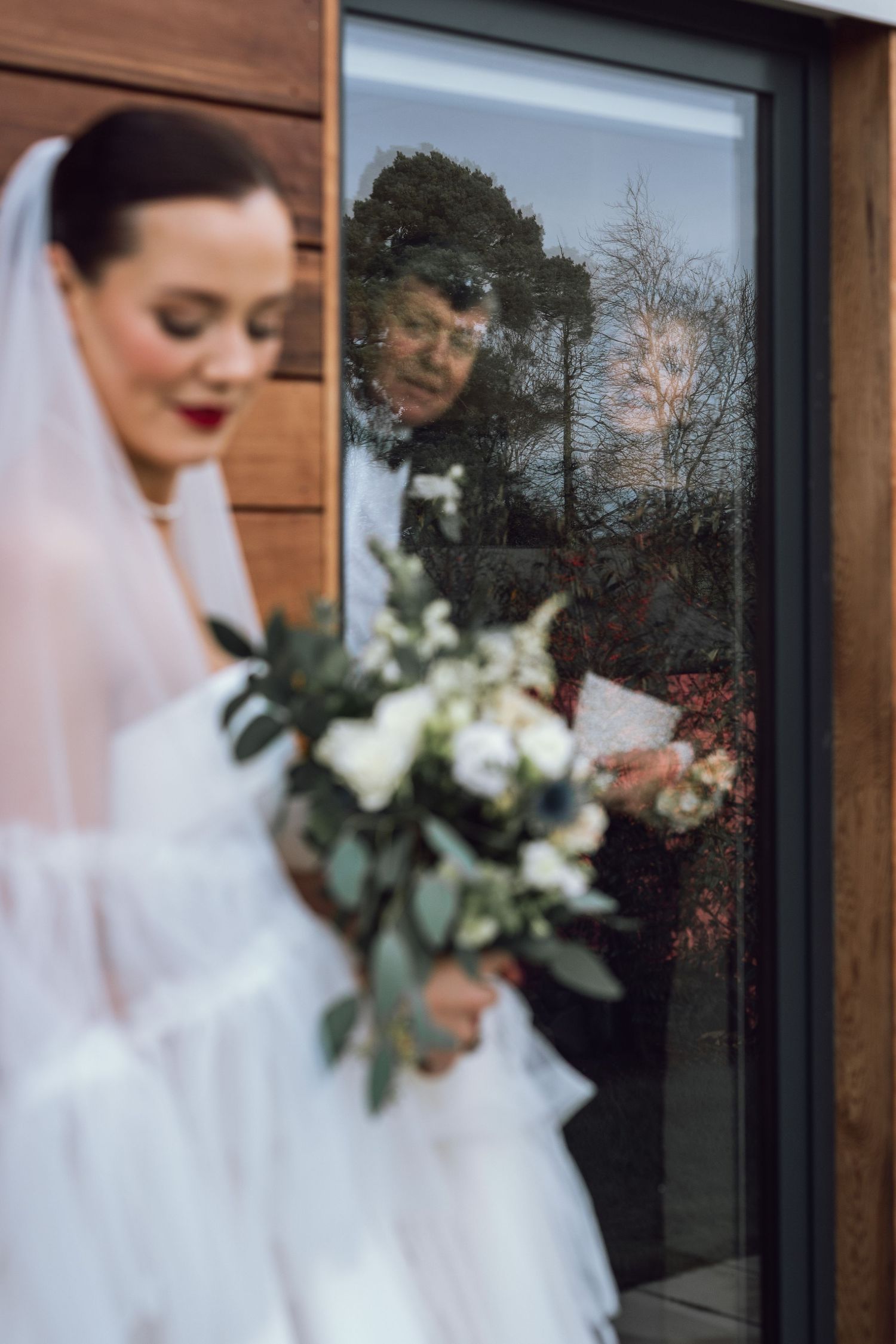 Bride holding white floral bouquet with guest reflected in glass door behind her.