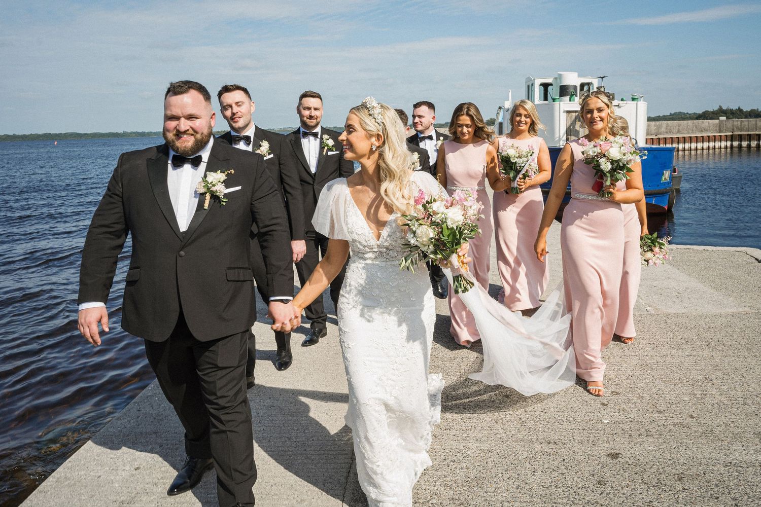 Bride and groom lead wedding party along waterfront promenade with bridesmaids in pink dresses.