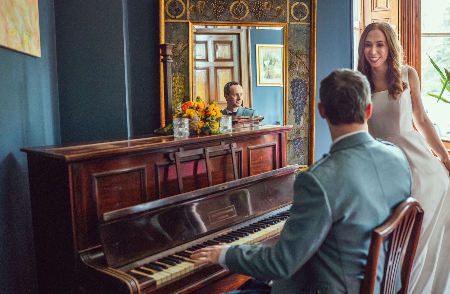 Bride smiling at groom playing piano in elegant blue room with ornate mirror and yellow flowers.