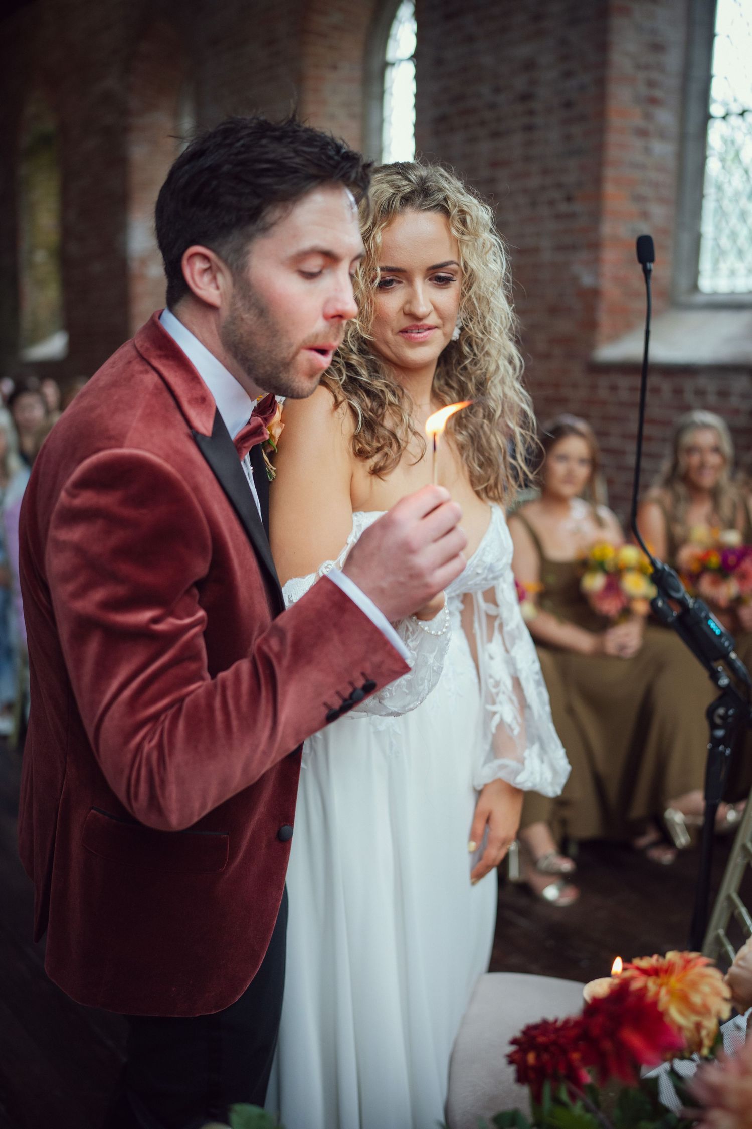 Groom in red velvet blazer lights unity candle with bride during wedding ceremony at red chapel Kells in Co Meath.