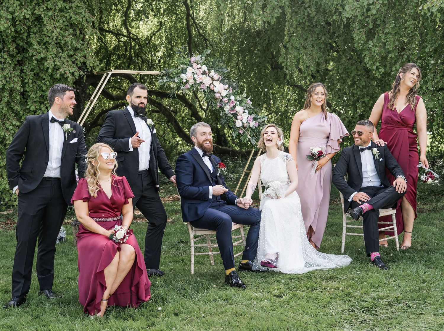 Wedding party poses casually on green lawn beside geometric floral arch with bridesmaids in burgundy and mauve gowns.
