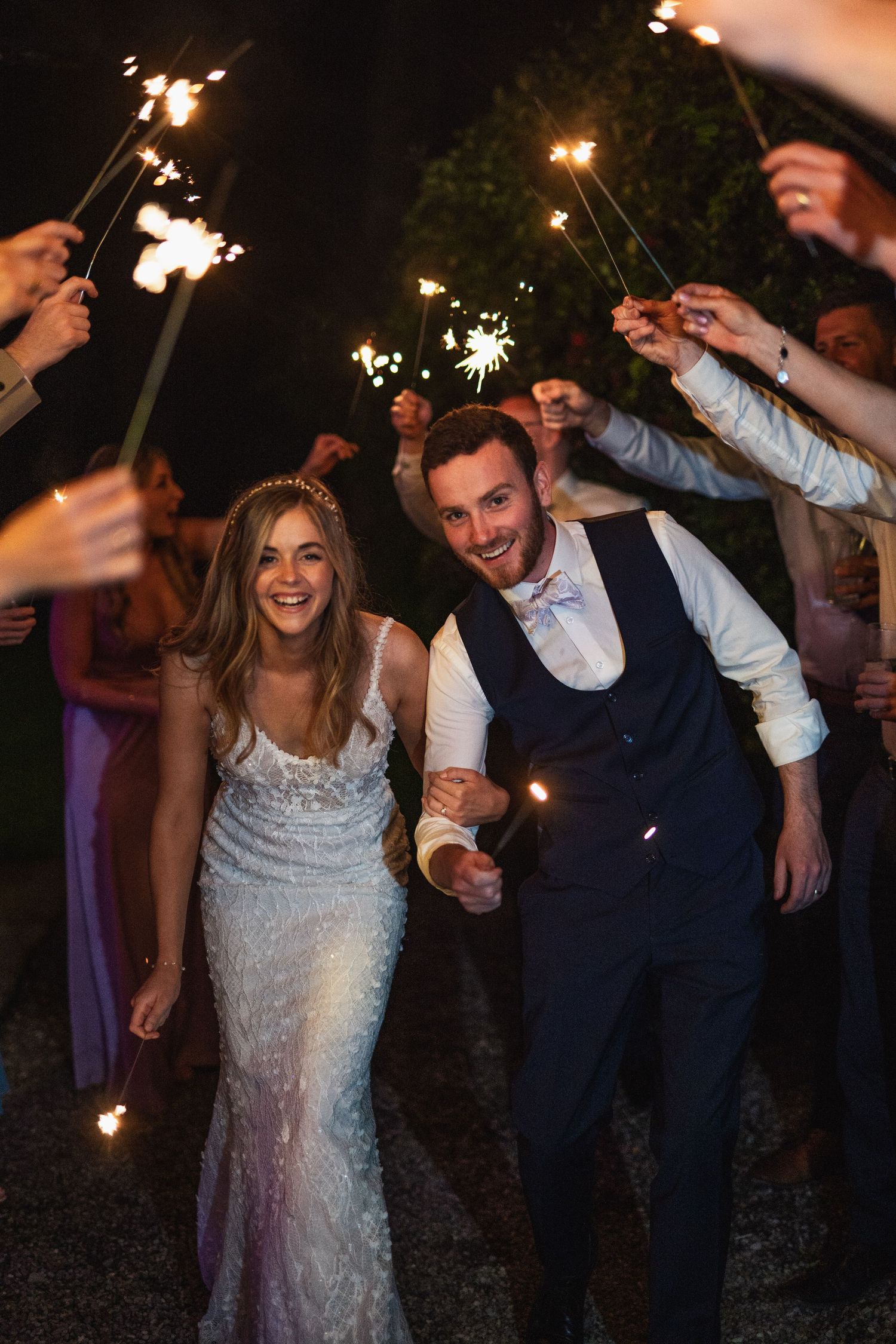 Joyful bride and groom walk through a tunnel of sparklers held by wedding guests at night.