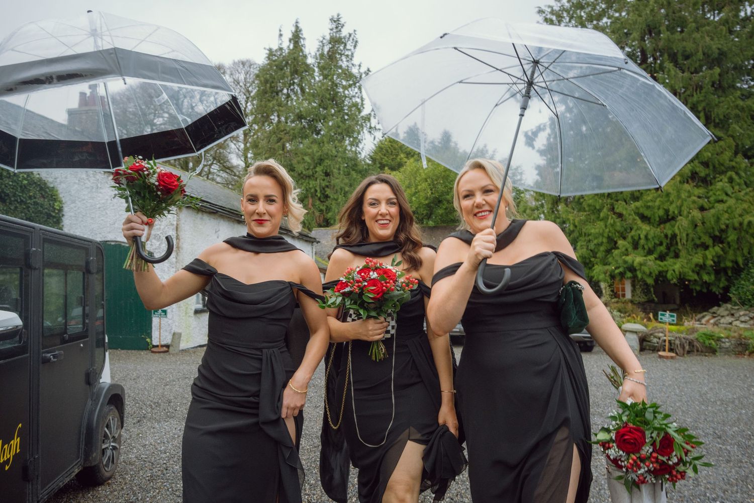 Three bridesmaids in black dresses holding red bouquets and umbrellas outdoors.