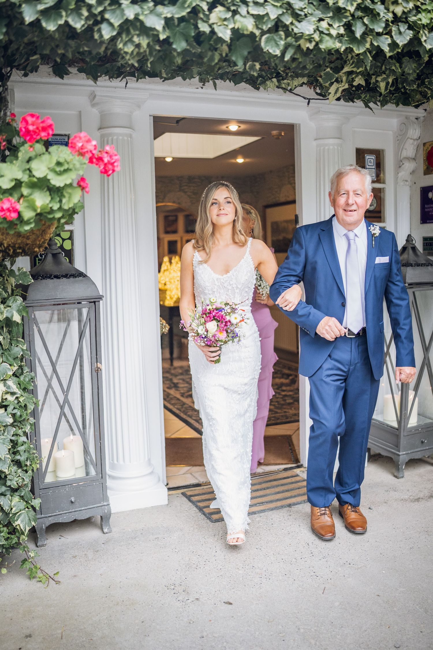 Bride in white lace gown walks arm in arm with father through ivy-covered venue entrance.