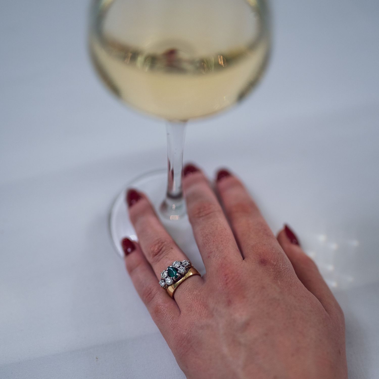Close-up of bride's hand with teal gemstone engagement ring and red nails beside a glass of white wine.
