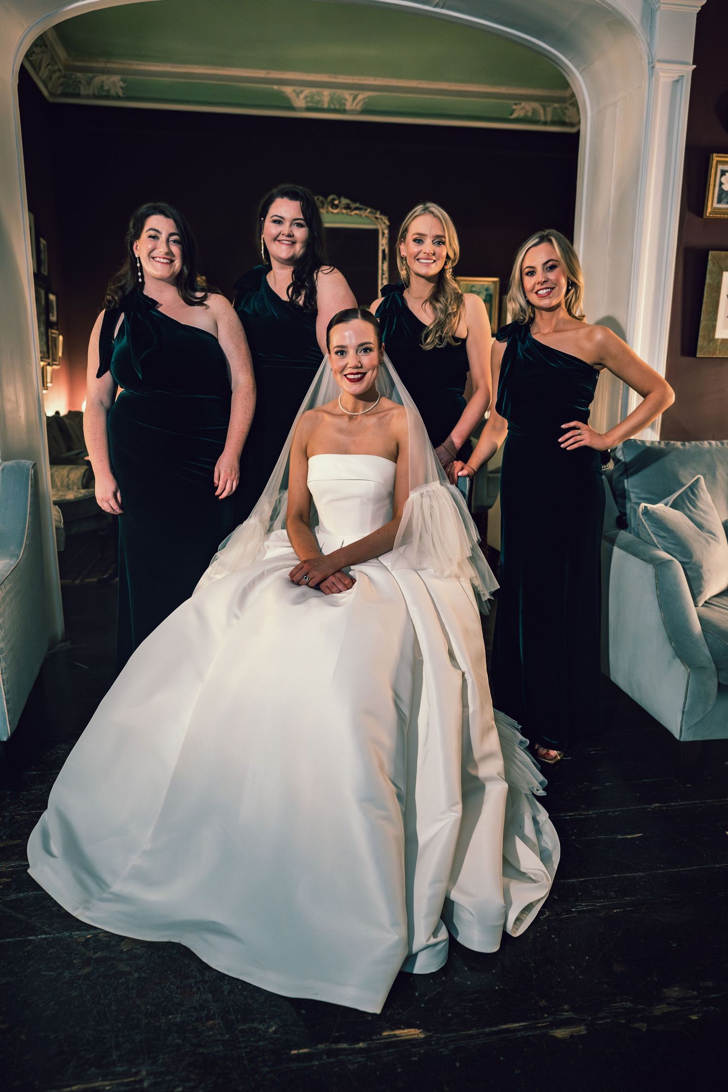 Bride in strapless white ball gown sits surrounded by four bridesmaids in elegant black one-shoulder dresses.