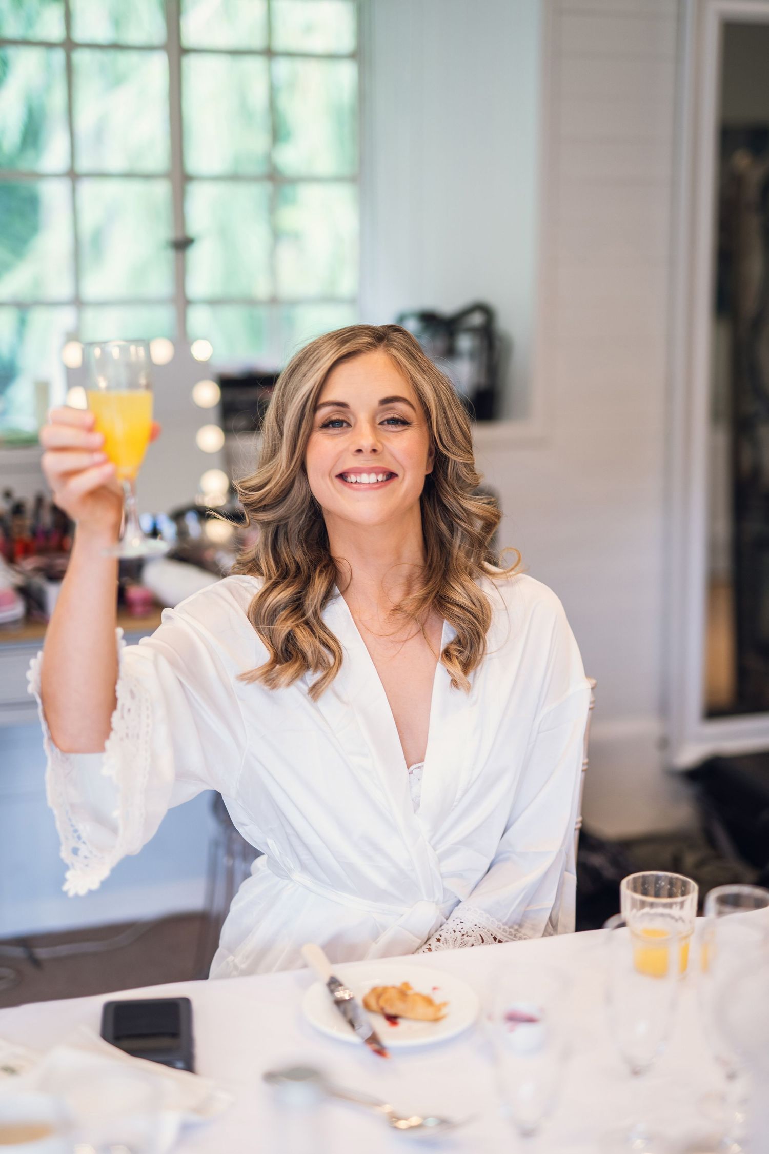 A smiling bride in a white robe raises a glass of orange juice during wedding morning preparations.