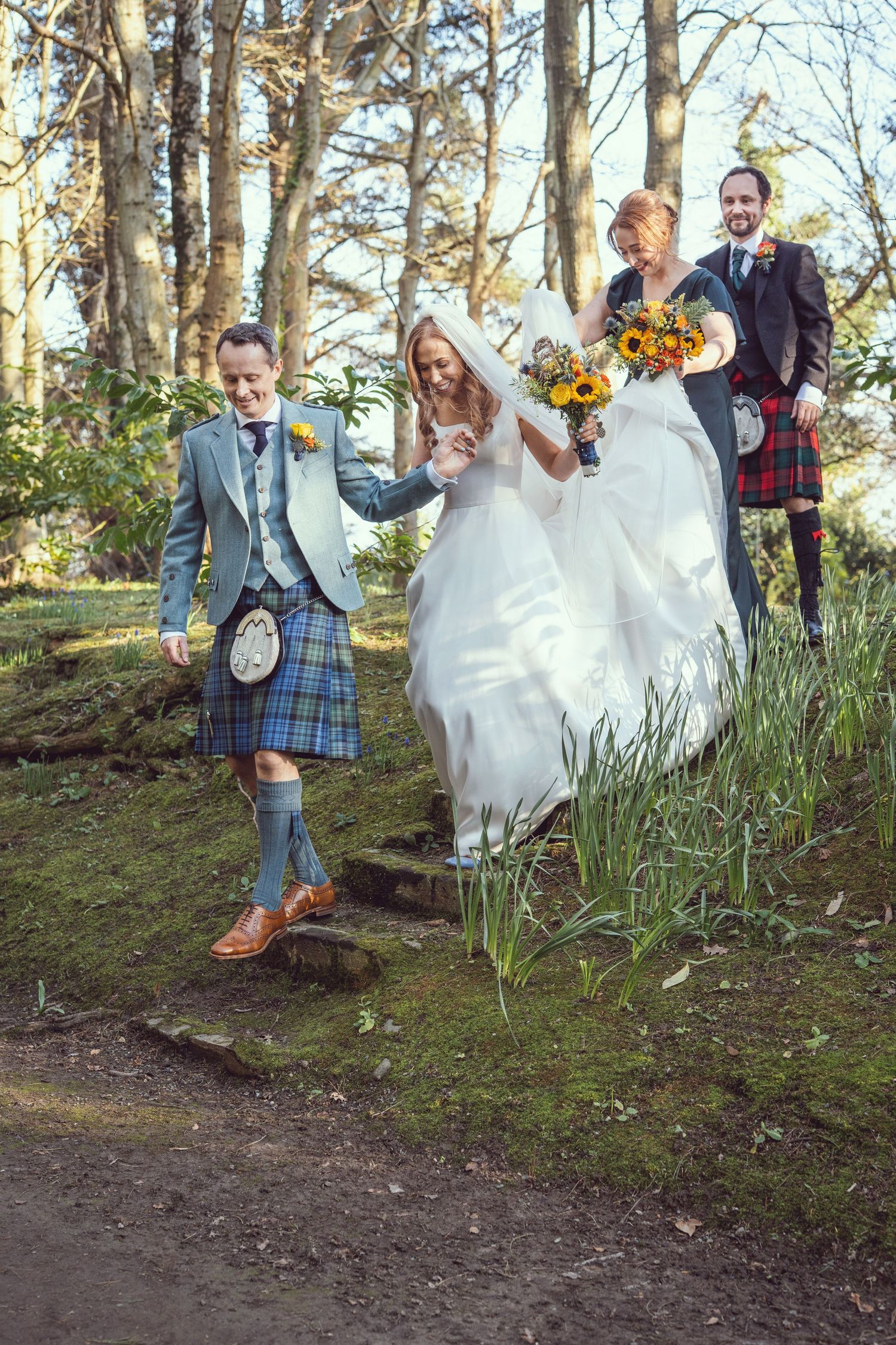 Wedding party with bride and groom in Scottish kilt descending stone steps in a sunlit woodland.