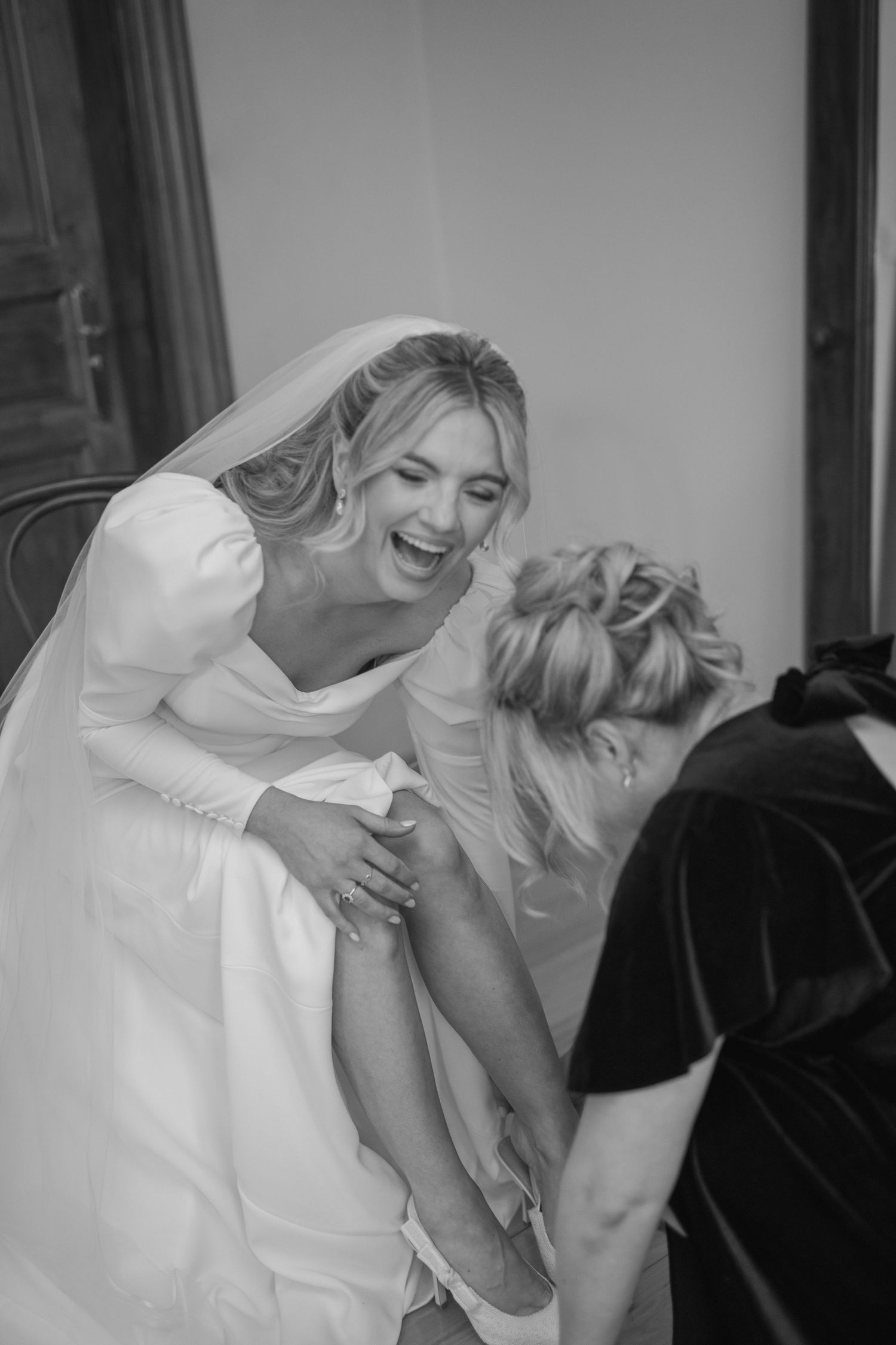 Laughing bride in white gown and veil gets assisted by bridesmaid during wedding preparations.