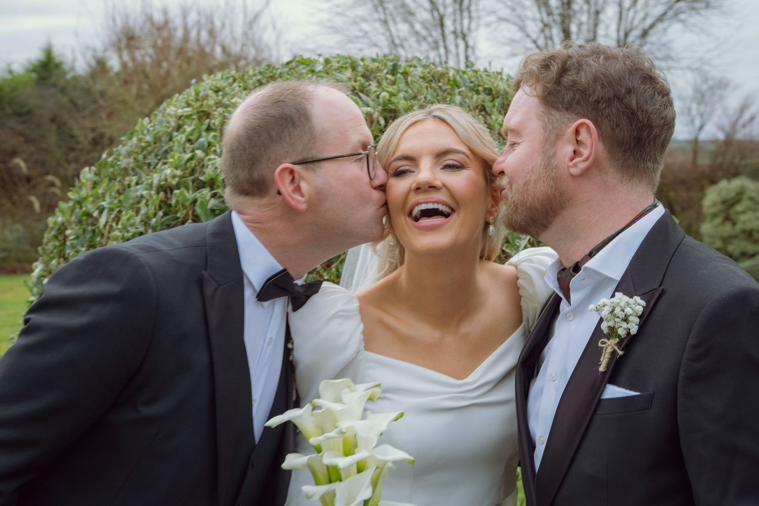Joyful bride laughs as two groomsmen in tuxedos kiss her cheeks outdoors.