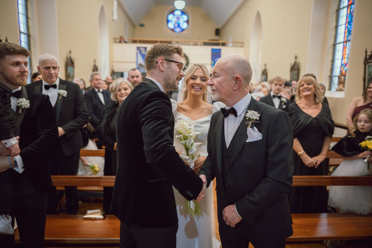 Groom shakes hands with older gentleman as bride looks on during church wedding ceremony.