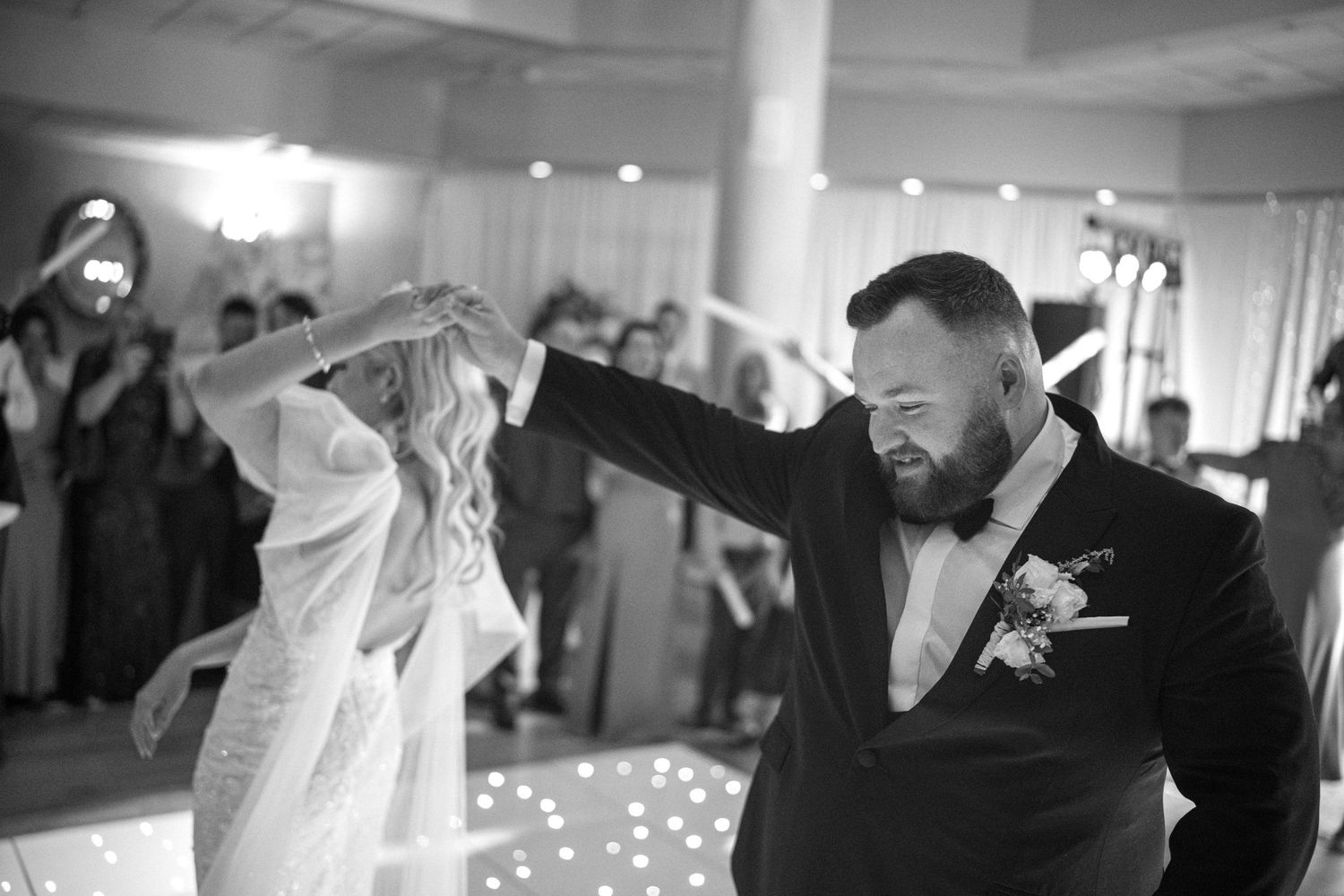 Groom in black suit twirling blonde bride on sparkling dance floor at wedding reception.
