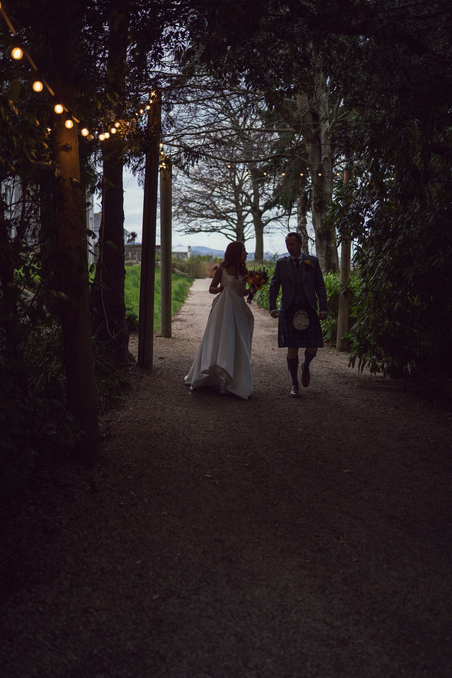Bride and groom in kilt walking hand-in-hand down a tree-lined path lit by warm festoon lights.