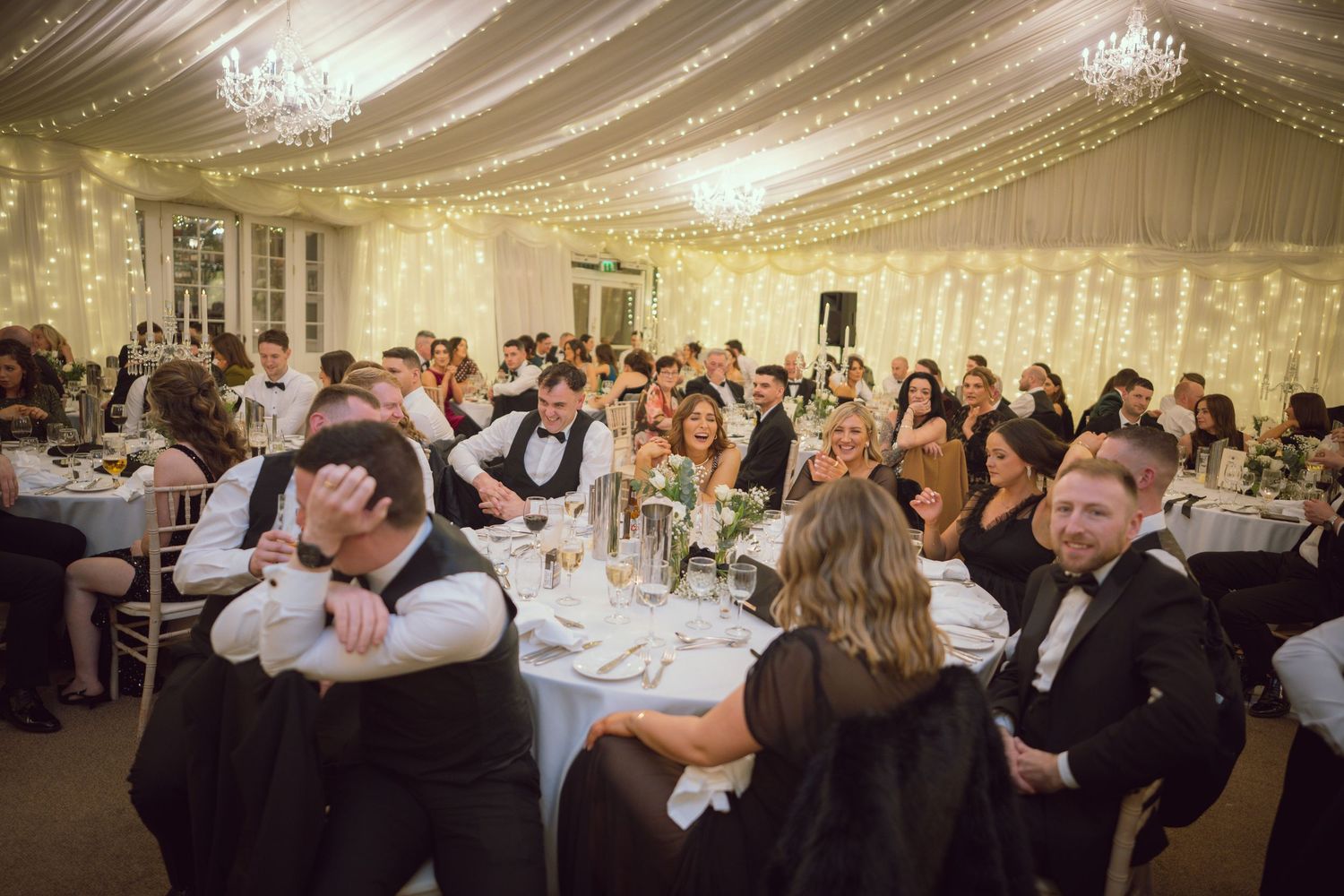 Elegant wedding reception in a marquee with fairy lights, guests laughing and celebrating at decorated tables.