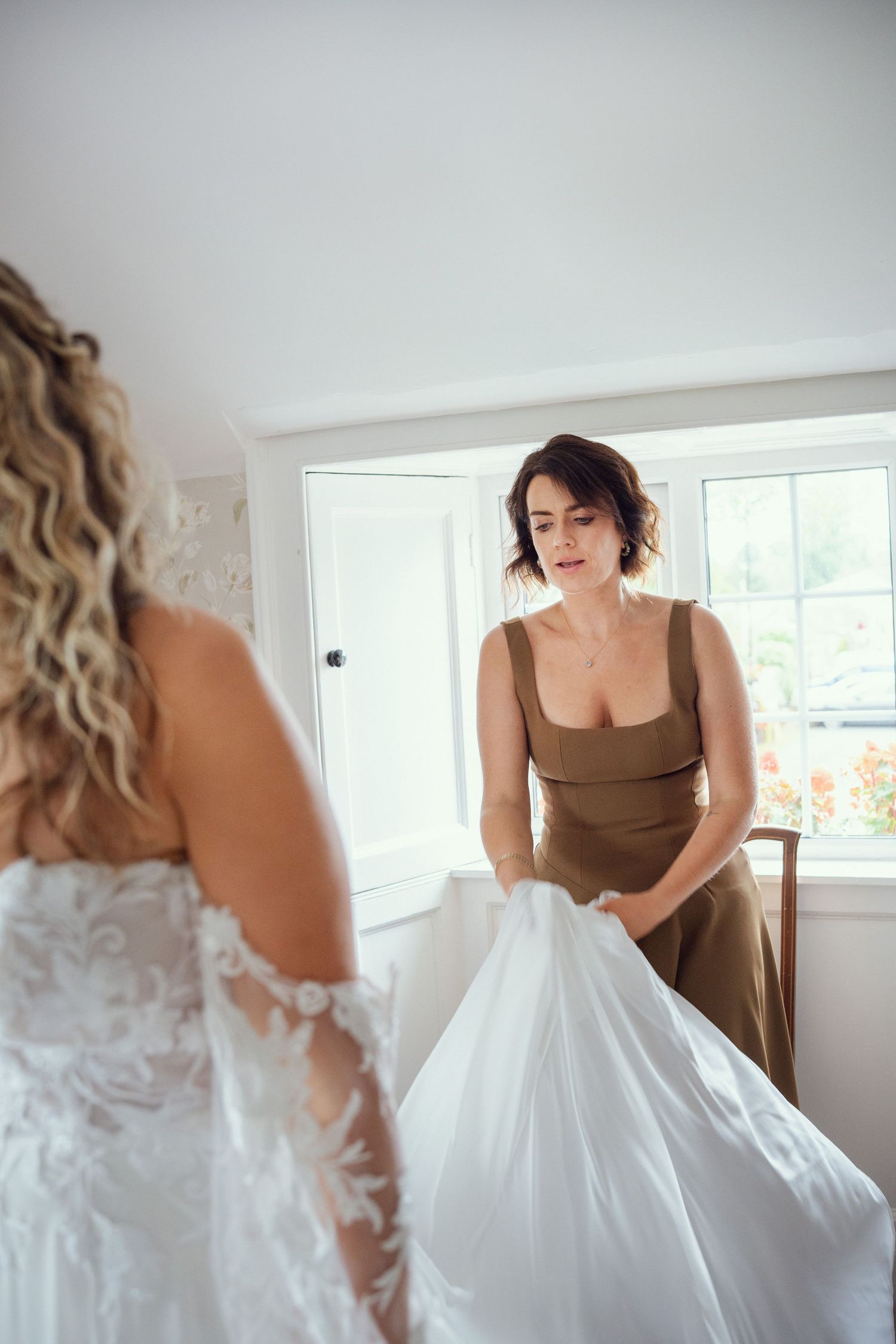 Bridesmaid in olive dress helping bride into her lace wedding gown near a bright window.