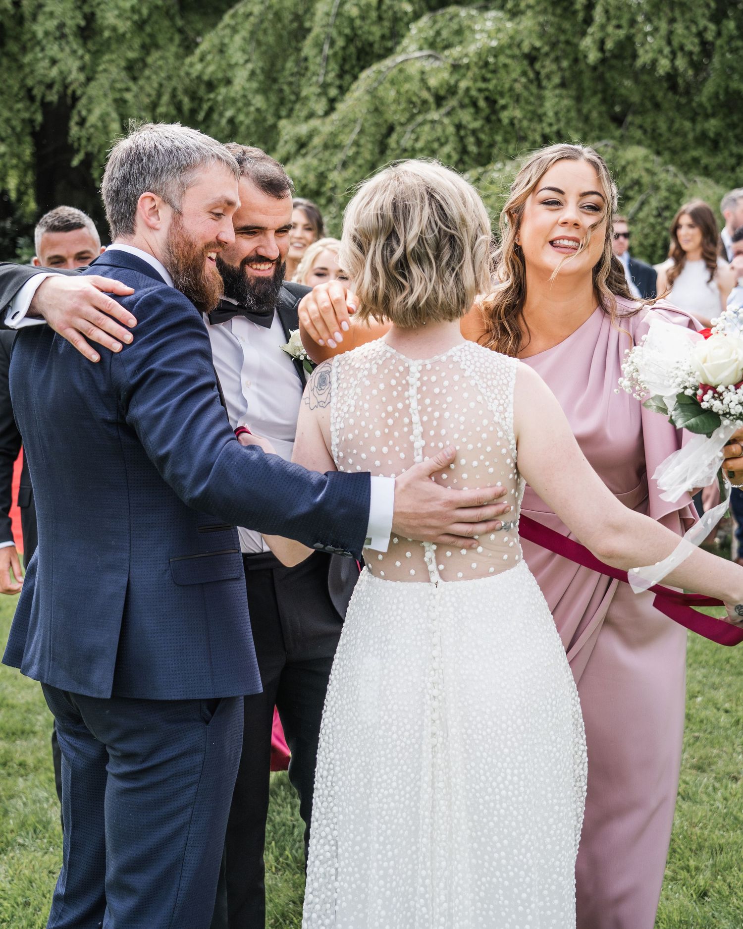 Bride in sheer polka-dot gown embraces guests while bridesmaid in pink holds bouquet at outdoor wedding celebration.