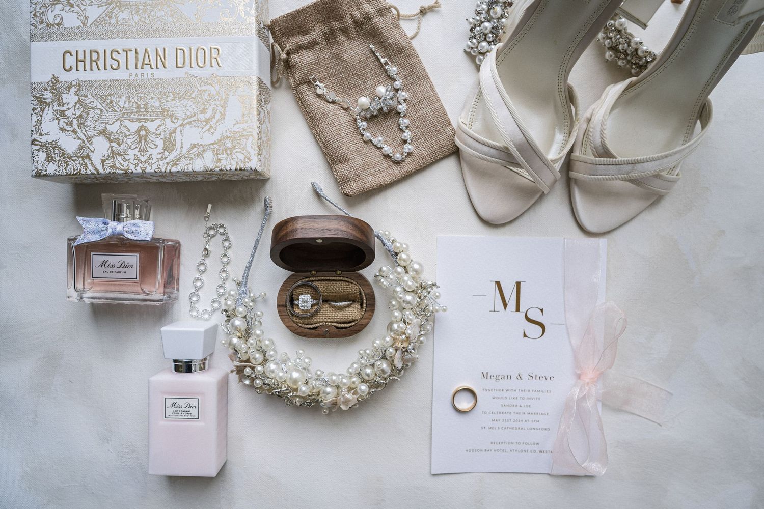 Luxury bridal flat lay featuring Christian Dior perfume, pearl jewelry, heels, and wedding invitation.