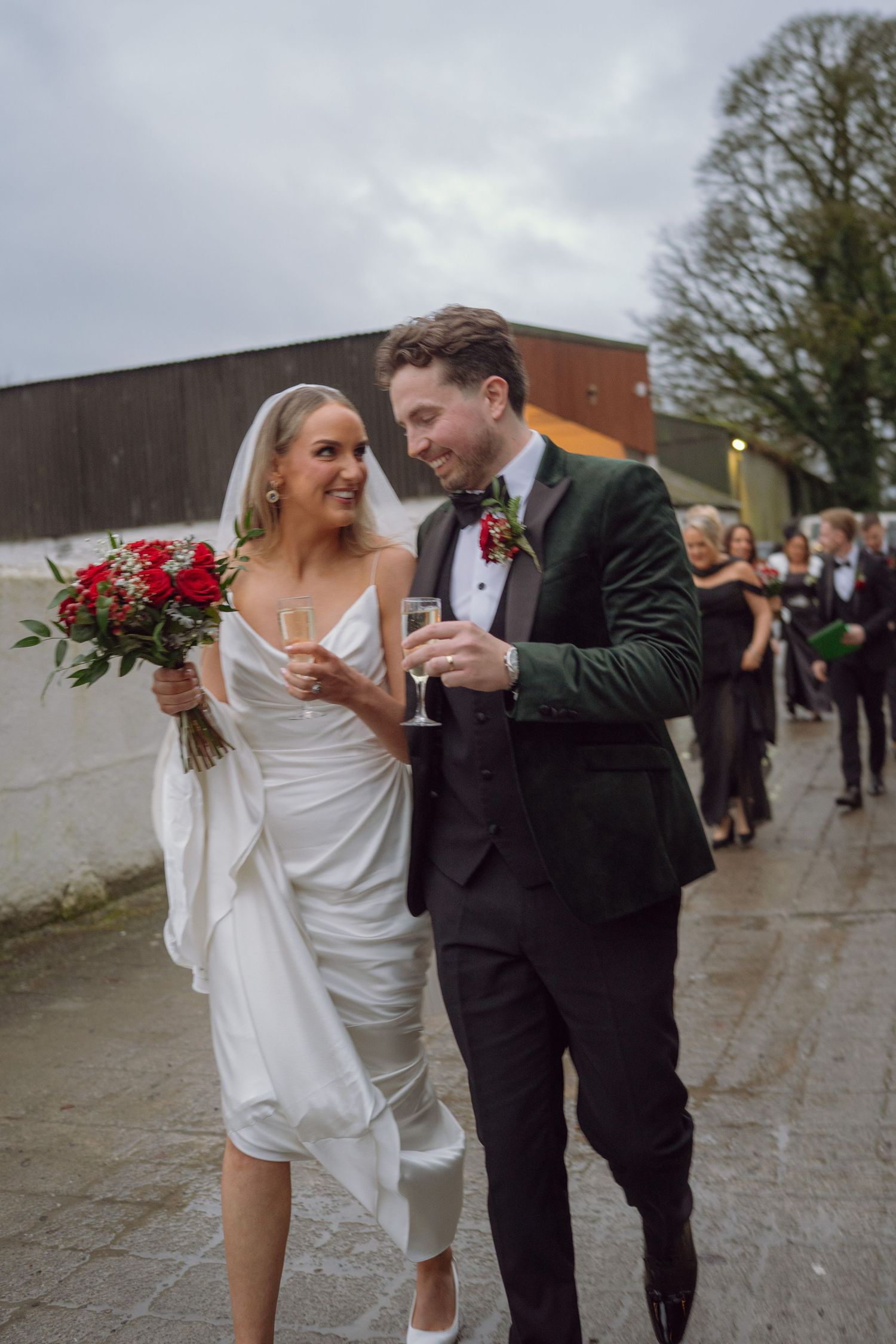 Bride and groom toast with champagne outside after wedding ceremony holding red bouquet.