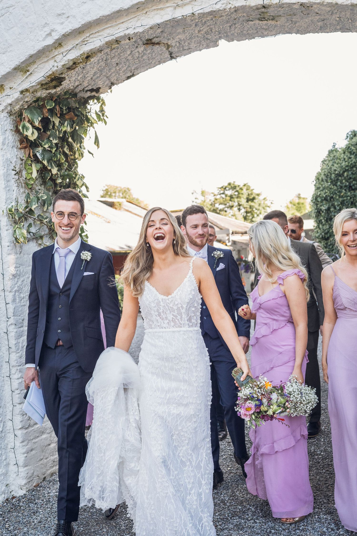 Laughing bride in beaded gown walks with groom and wedding party through a stone archway.