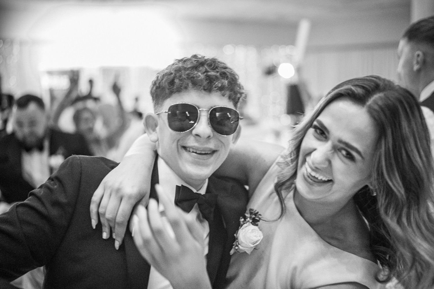 Young man in sunglasses and bow tie posing with laughing bride in black and white wedding reception photo.