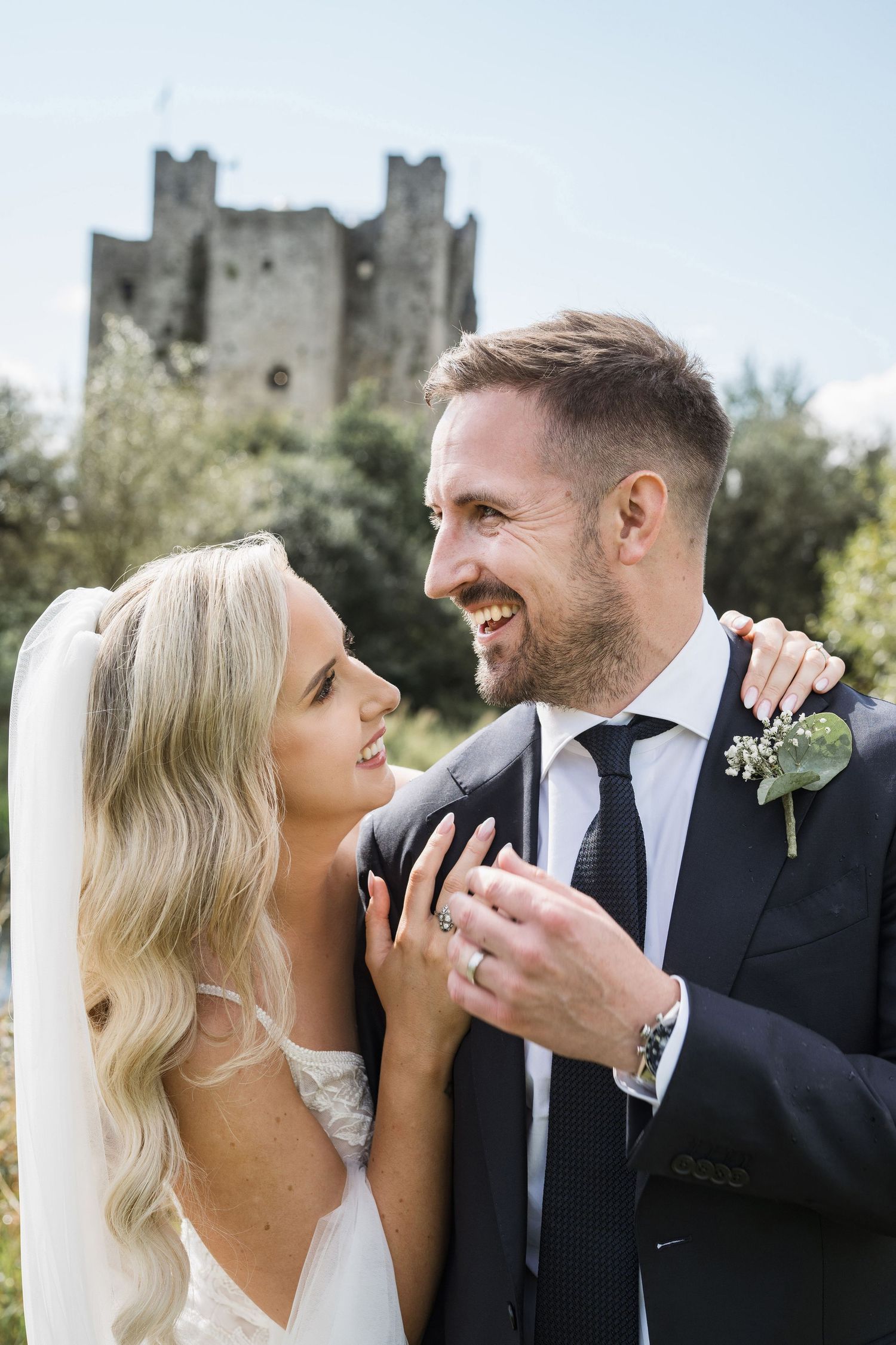 Smiling bride and groom pose together with a historic stone castle in the background on their wedding day.