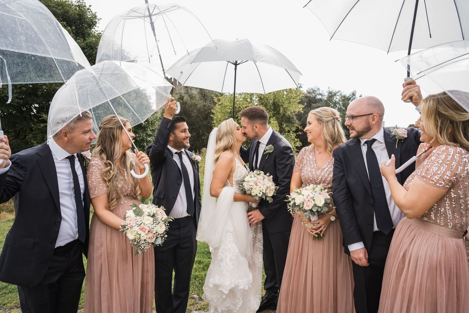 Wedding party with bride and groom kissing surrounded by bridesmaids in pink dresses holding clear umbrellas outdoors.