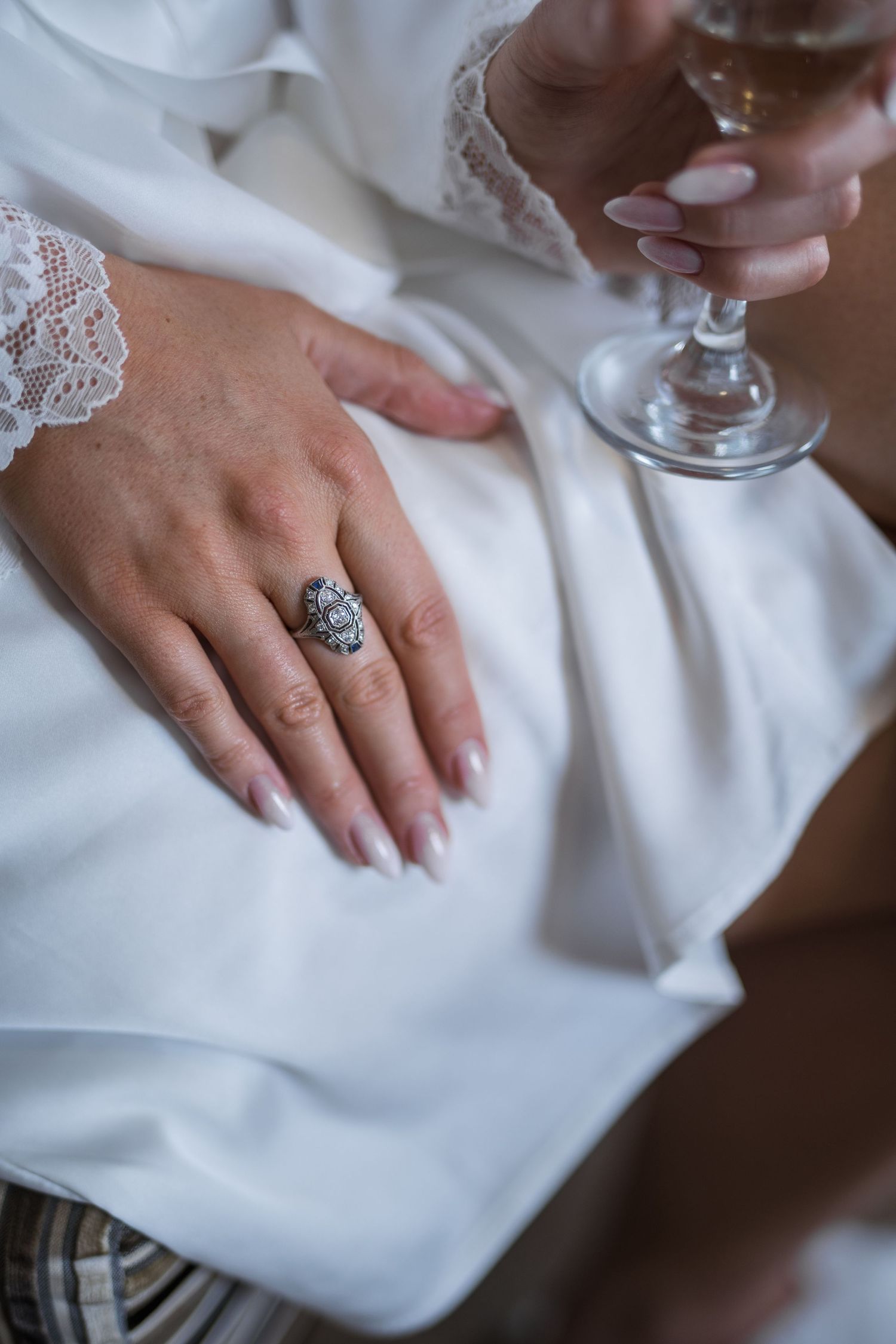 Close-up of bride's hand wearing ornate vintage ring while holding champagne glass in white robe.