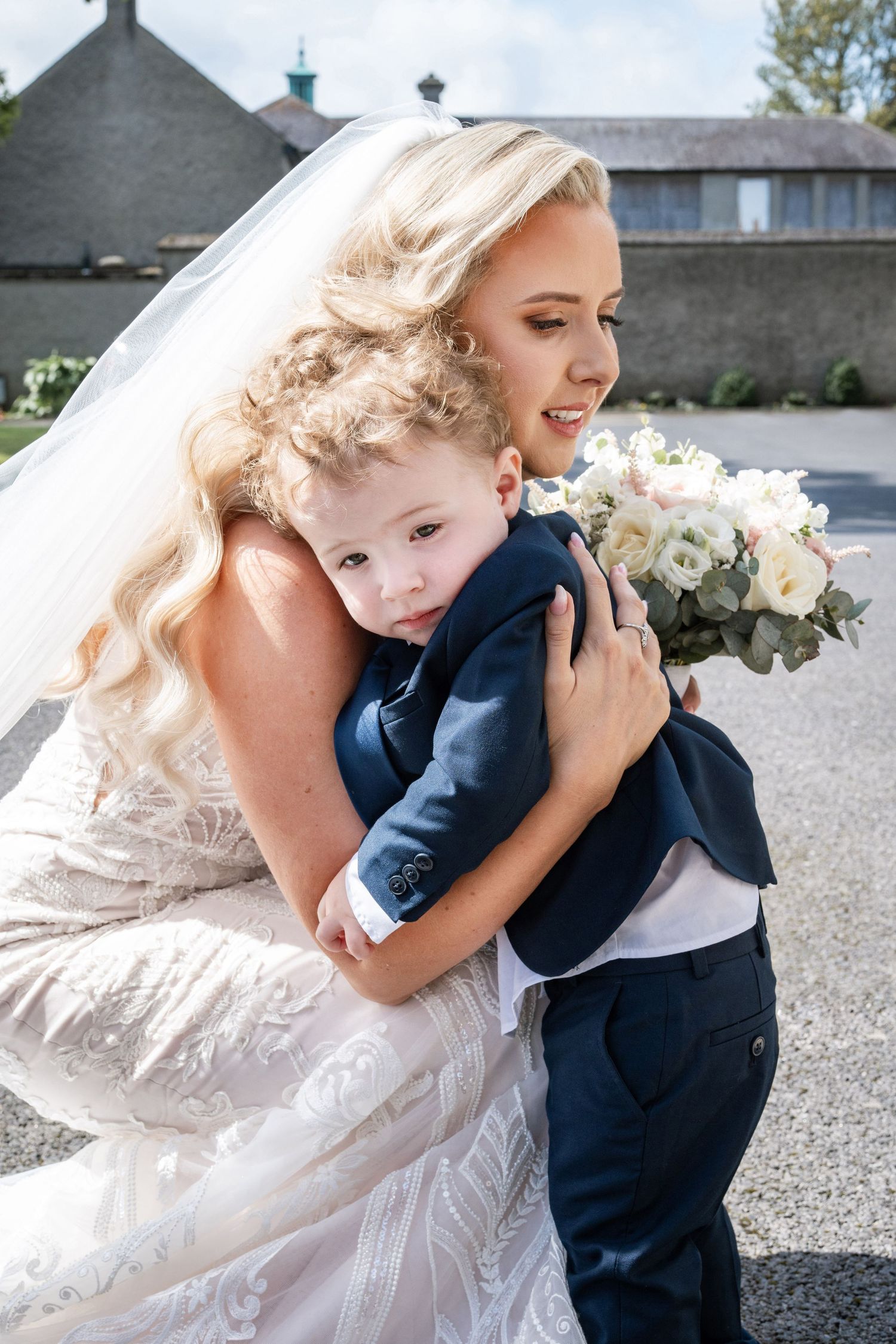 Blonde bride in lace gown tenderly hugging a young boy in a navy suit outdoors.