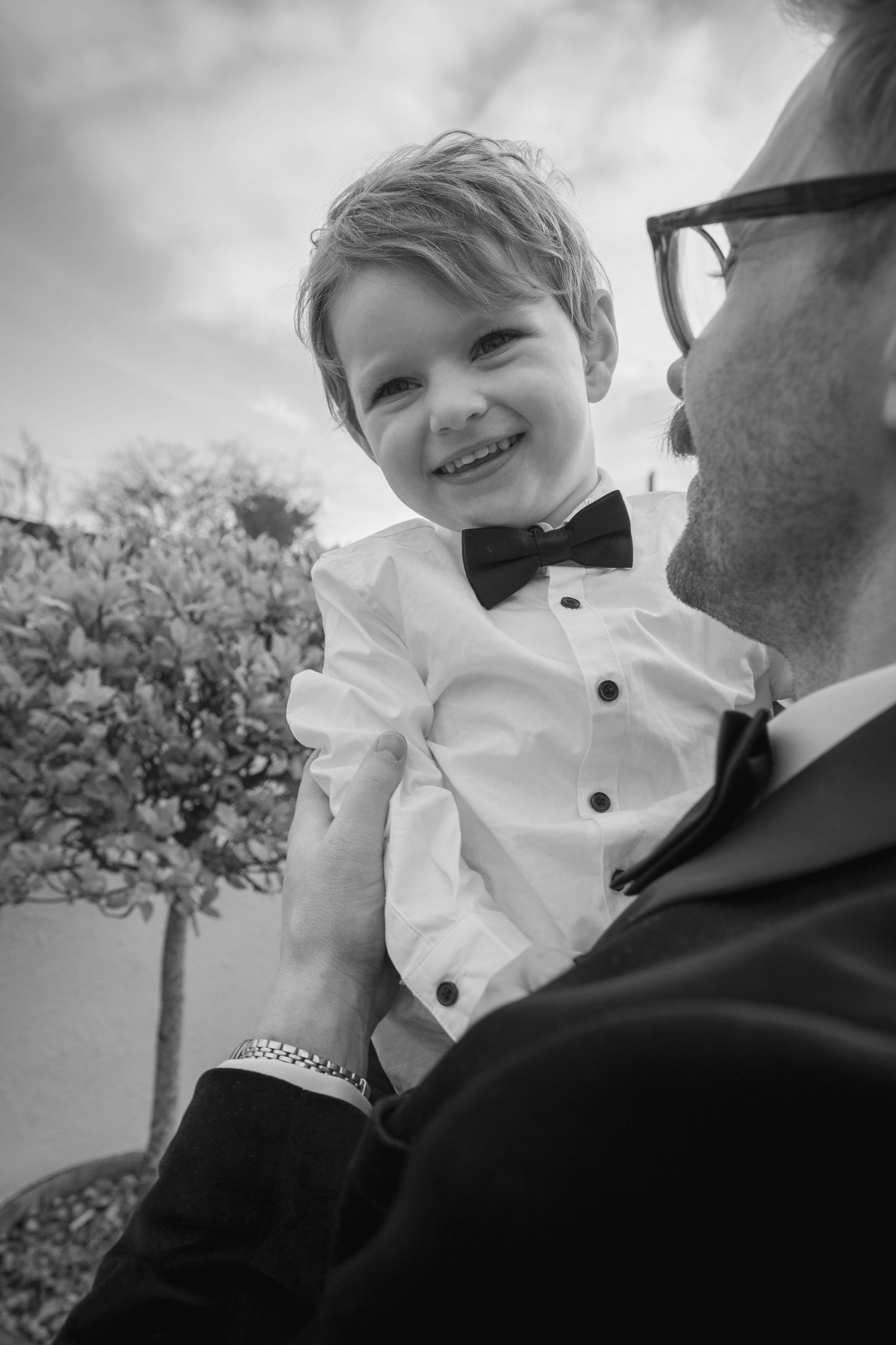 Smiling young boy in white shirt and bow tie being held by a suited groom in black and white photo.
