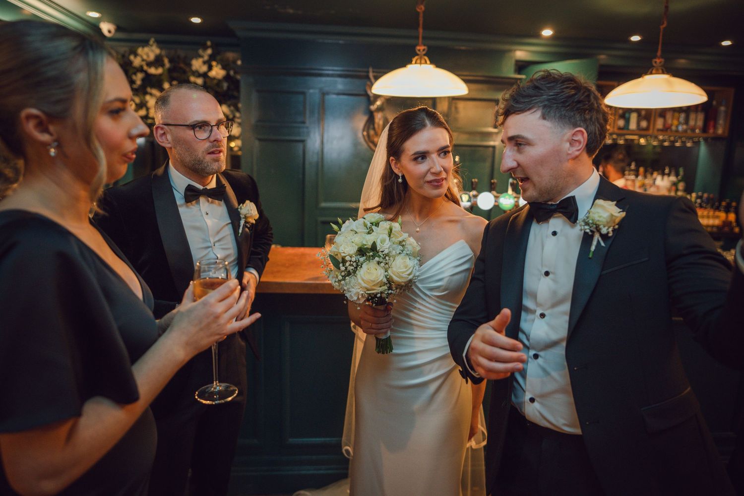 Bride in white gown holding bouquet chats with guests in tuxedos at elegant wedding reception bar at Mount Juliet wedding in Kilkenny.