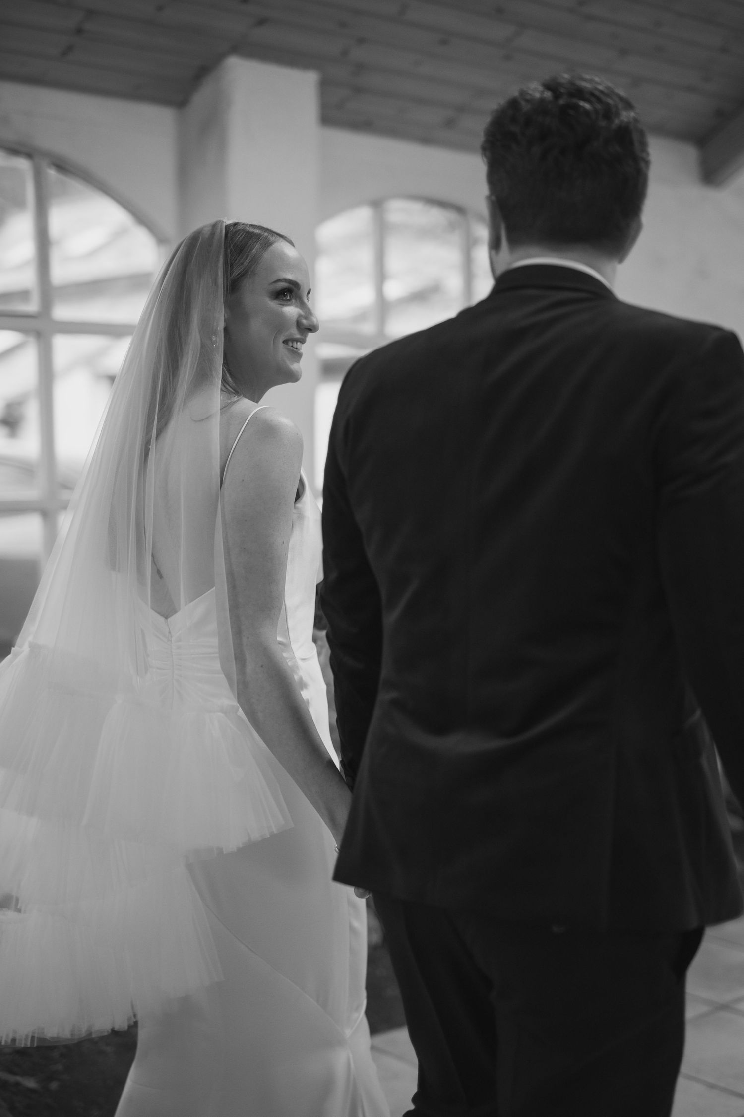 Bride in white gown smiling at groom in black and white photo.