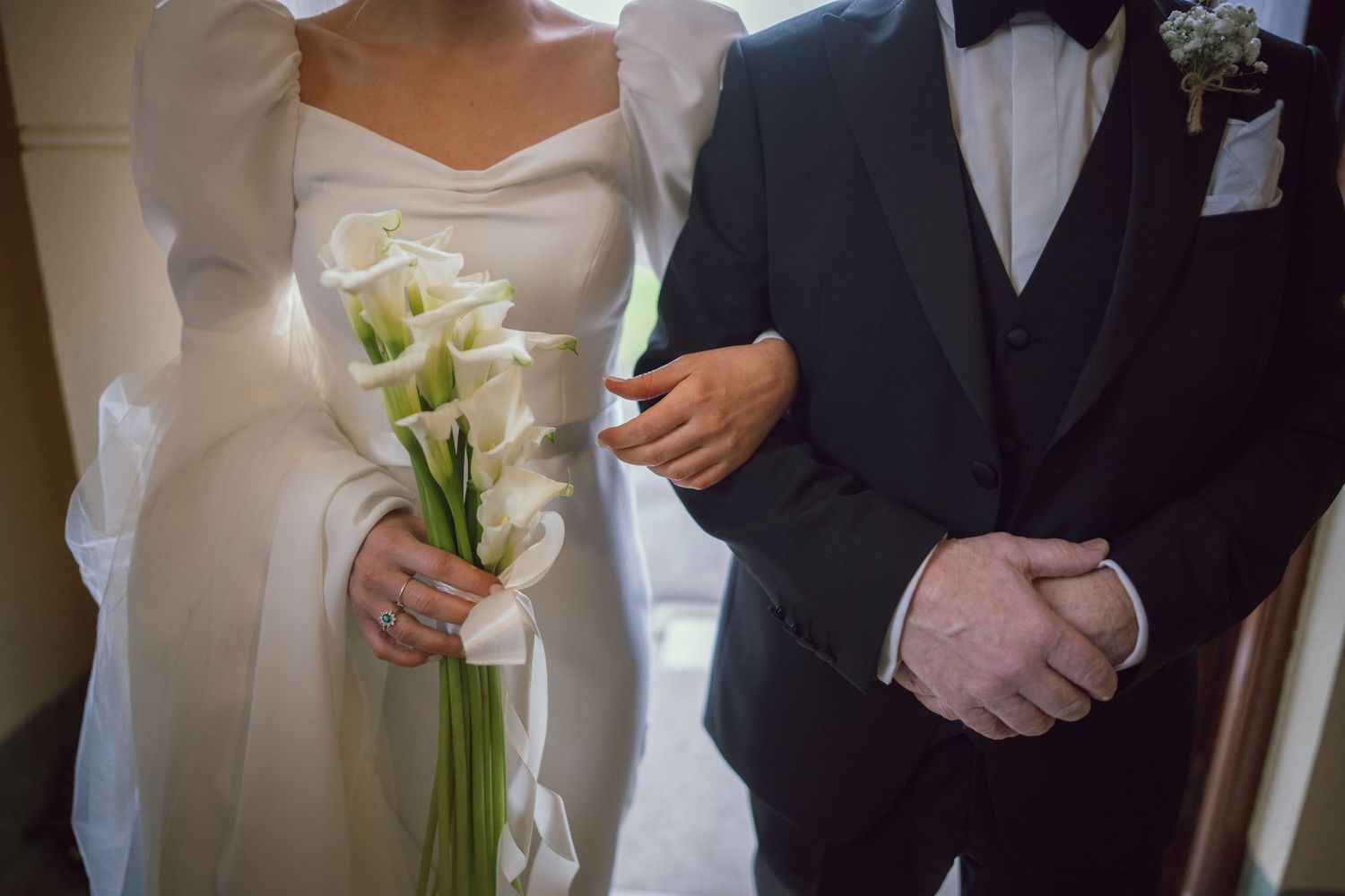 Close-up of bride holding white calla lily bouquet while standing beside groom in black tuxedo at wedding.