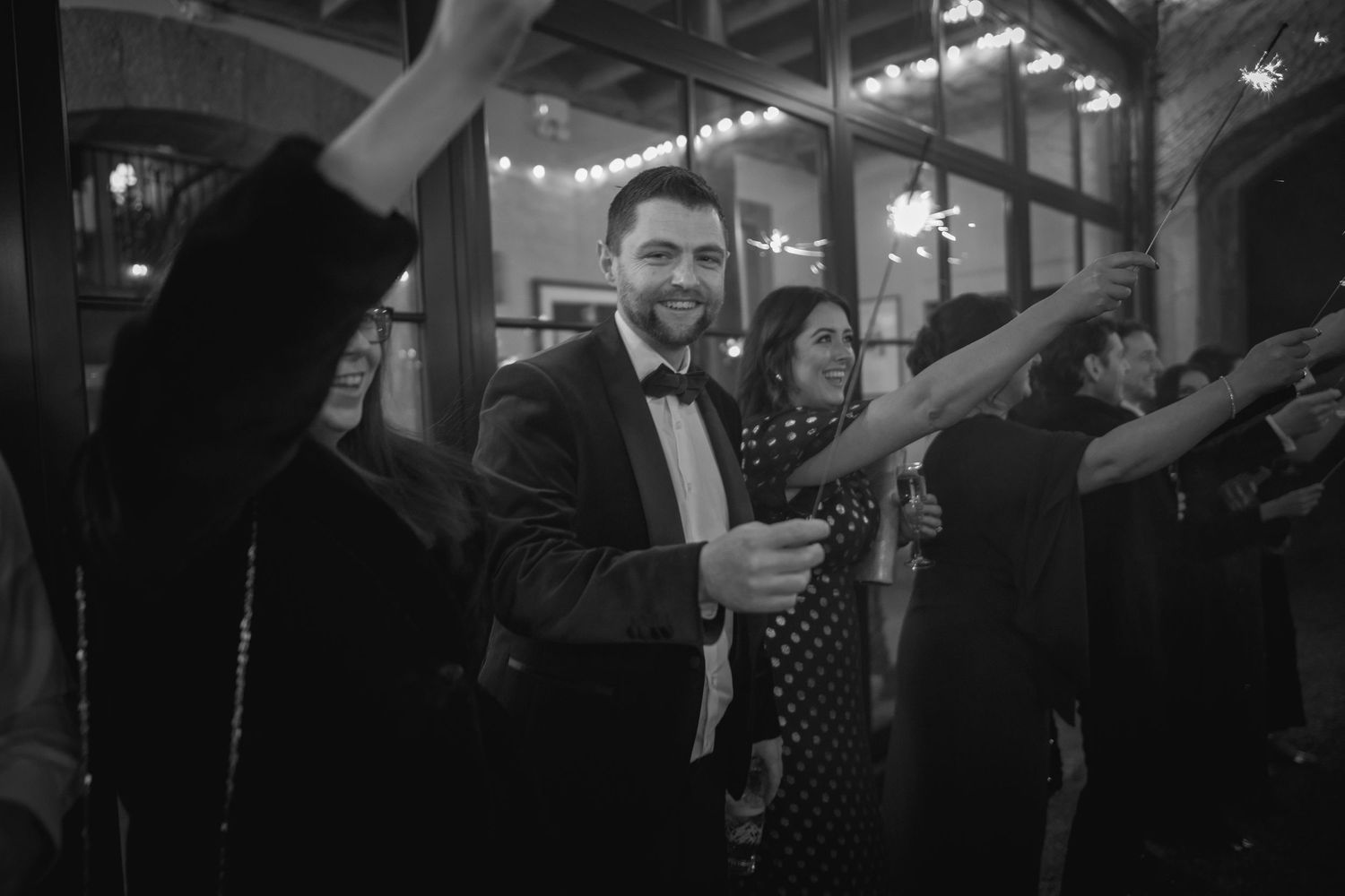 Smiling groom in tuxedo surrounded by guests holding sparklers at wedding reception, black and white.