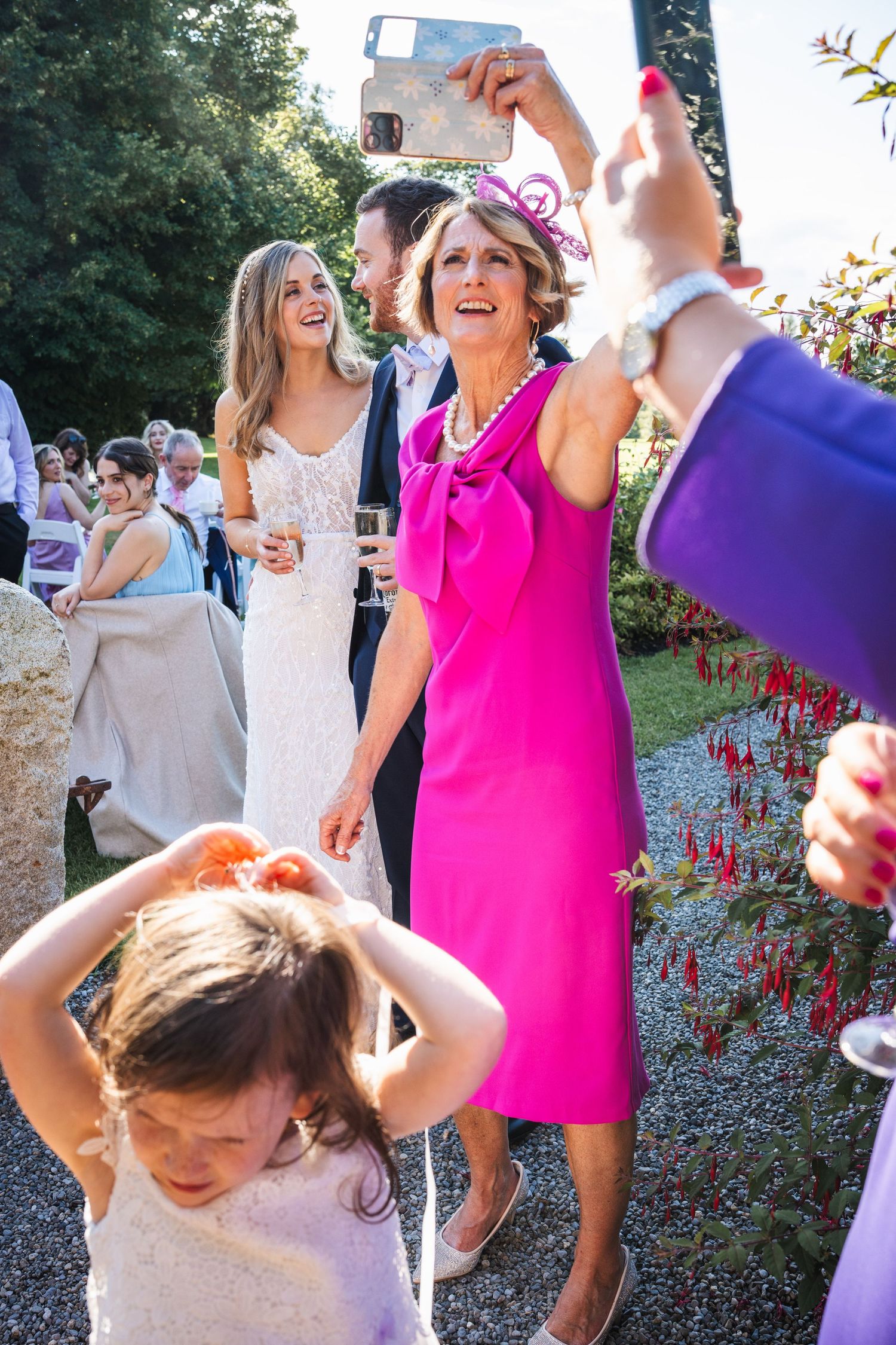 Wedding guest in pink dress taking selfie at lively outdoor reception.