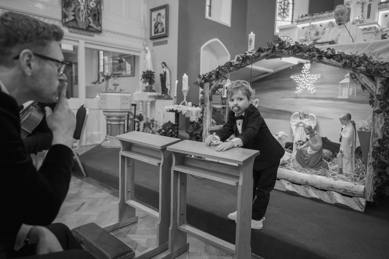 A young boy in a bow tie leans on a desk near a nativity scene inside a decorated church.
