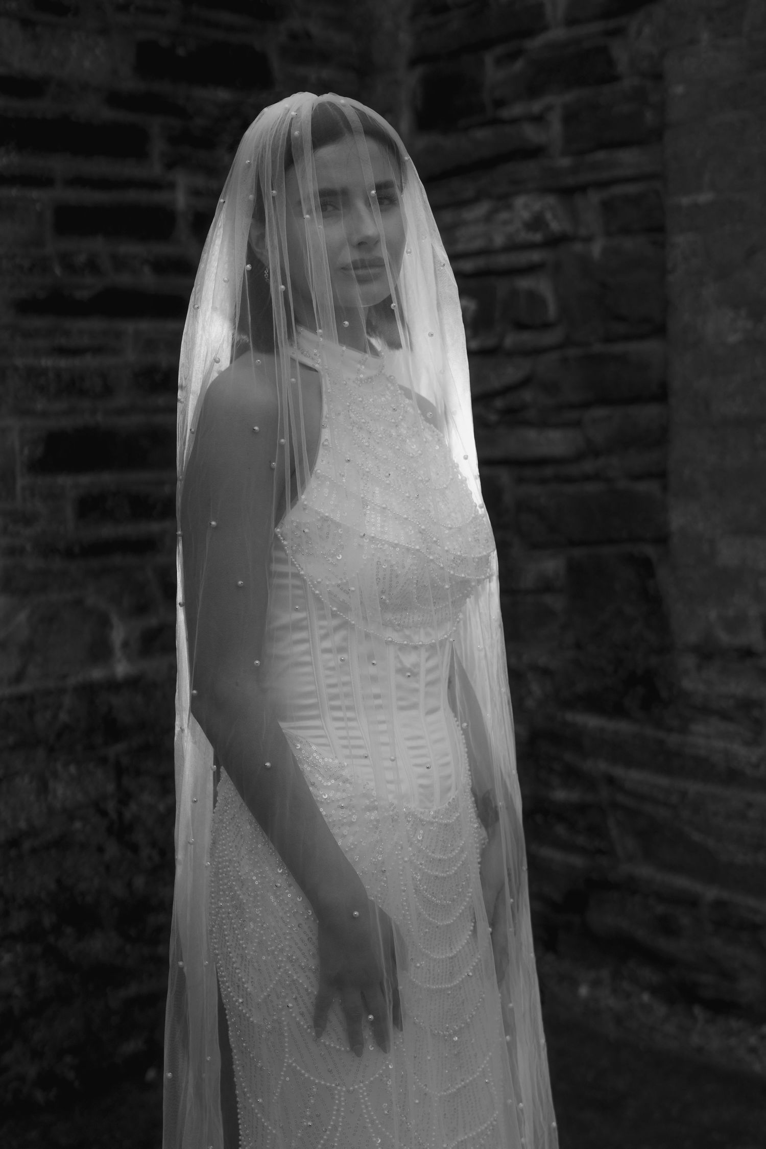 Black and white portrait of a bride in a lace gown and pearl-adorned veil against a stone wall backdrop.